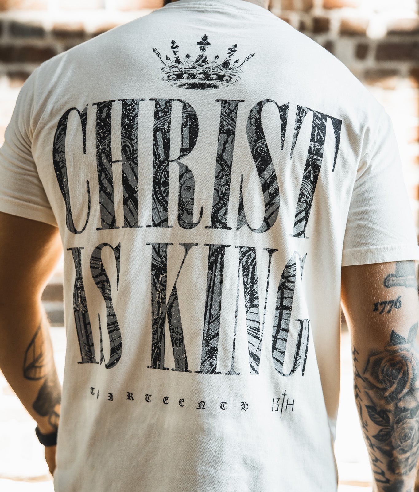 A person wearing the Christ is King Premium Tee - Ivory by 13th, featuring bold "CHRIST IS KING" text, a crown graphic above, and "thirteen 13th" below. Visible arm tattoos complement this standout Christian shirt.