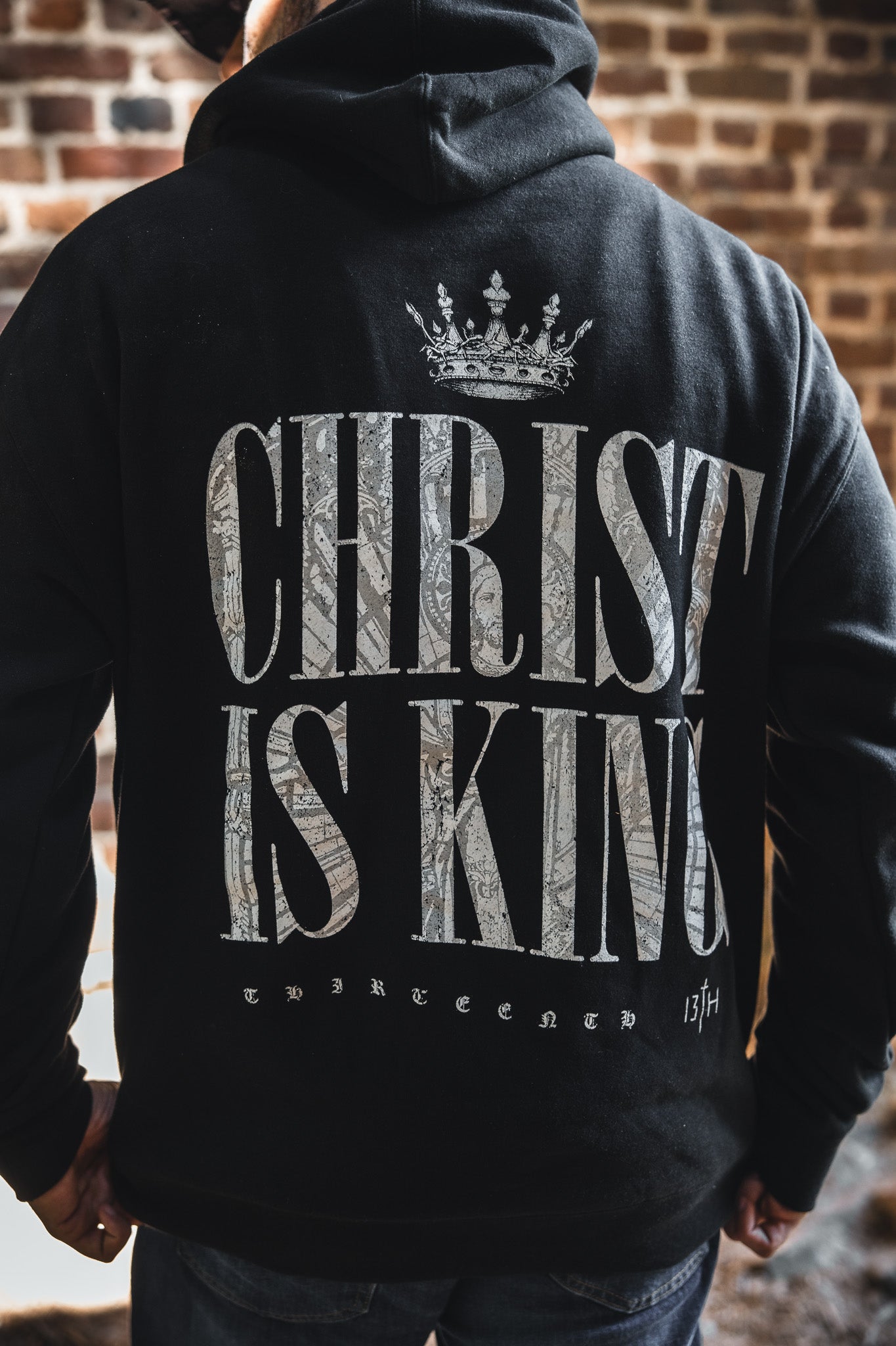 Someone wears the Christ is King Premium Hoodie by 13th: a black Christian apparel piece with bold "CHRIST IS KING" on the back, a crown graphic above, and "THIRTEEN 13TH" below—a powerful faith statement.