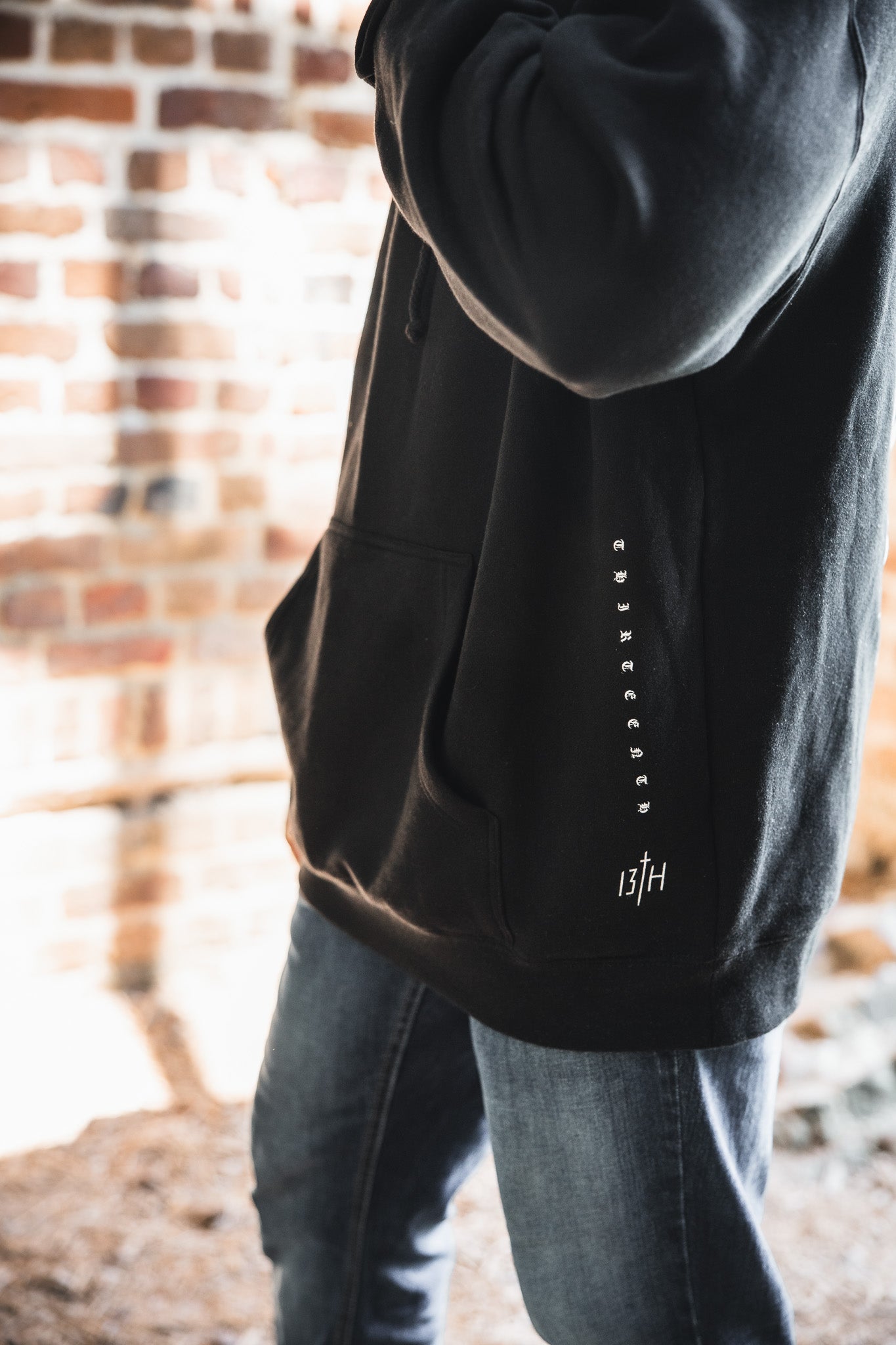 Wearing the 13th brand Christ is King Premium Hoodie in black with white side text and a "13TH" logo near the pocket, paired with blue jeans, a person stands in front of a sunlit brick wall.