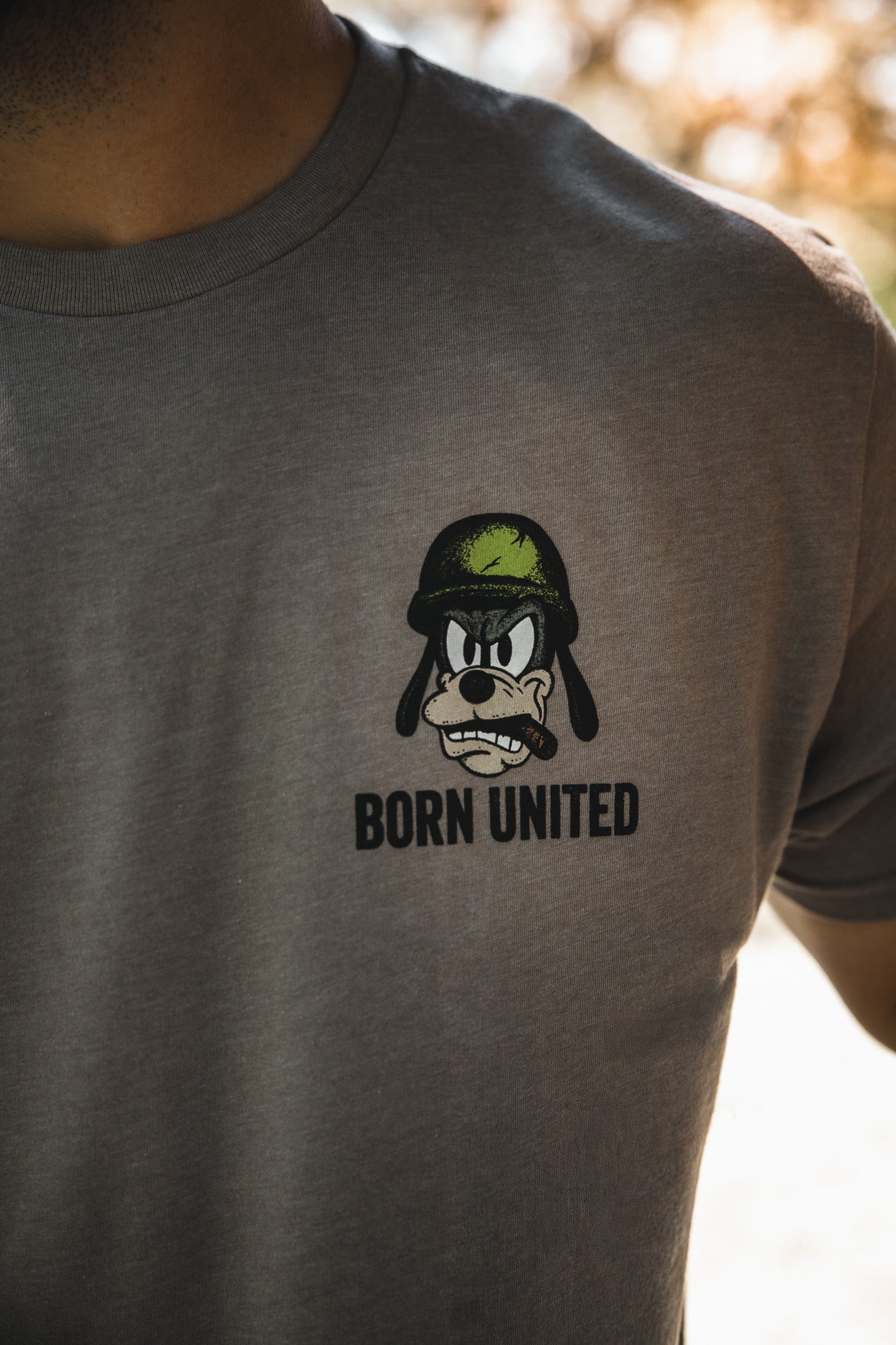 A person wears the Ain't Goofin' Around gray T-shirt by Born United Private Label, featuring a cartoon dog in a green army helmet above "BORN UNITED" and "Ain't Goofin' Around." The photo highlights the shirt’s design; the face is partly out of frame.