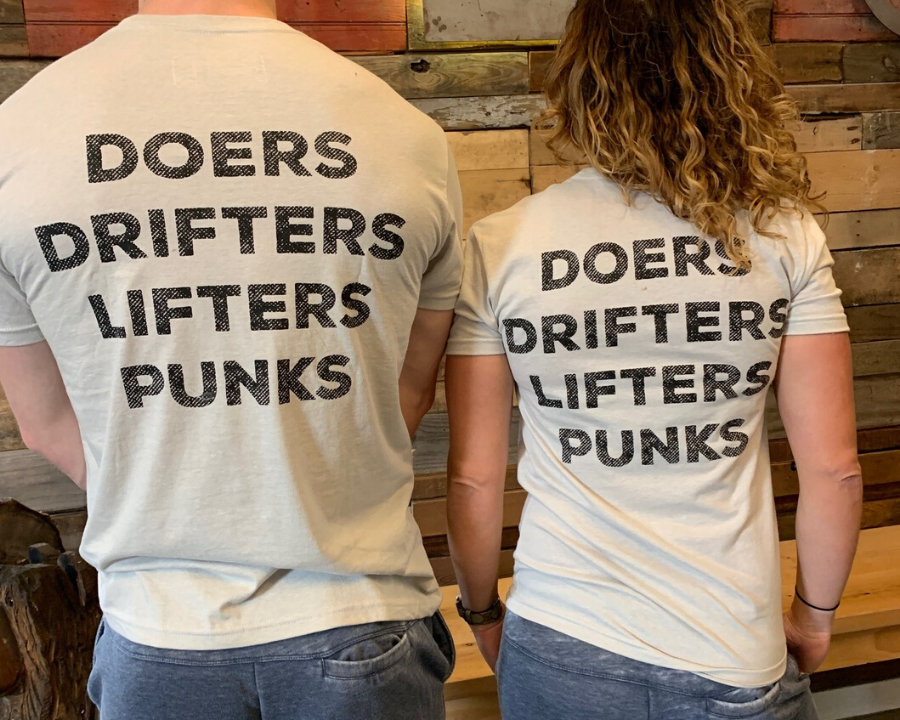Two people, backs to the camera, wear the Doers Drifters Lifters Punks Unisex Tee by Tango Charlie Apparel. The bold slogan stands out against light fabric as they rep their crew in front of a rustic wooden wall.
