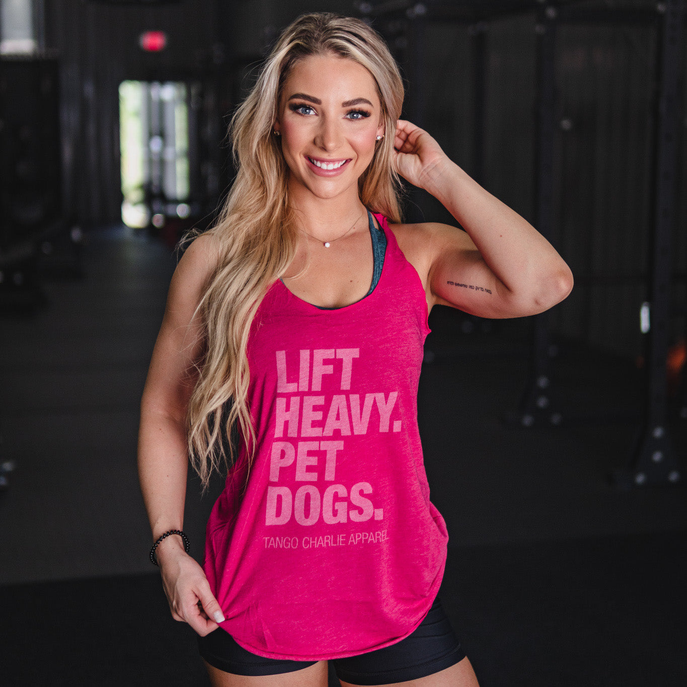 A smiling woman with long blonde hair stands in a dark, blurred gym, wearing the Lift Heavy. Pet Dogs.- Womens Pink Tank by Tango Charlie Apparel and black shorts. Her left hand touches her hair.