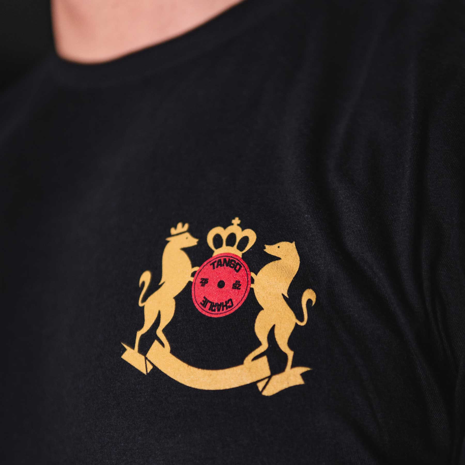 Close-up of the Deadlift Addiction - Tee by Tango Charlie Apparel, a black T-shirt with a gold emblem showing two stylized animals and a crown around a red circle featuring symbols and TANEO JAPAN. Perfect for anyone who takes the lifter generals warning seriously, its as essential as your next deadlift.