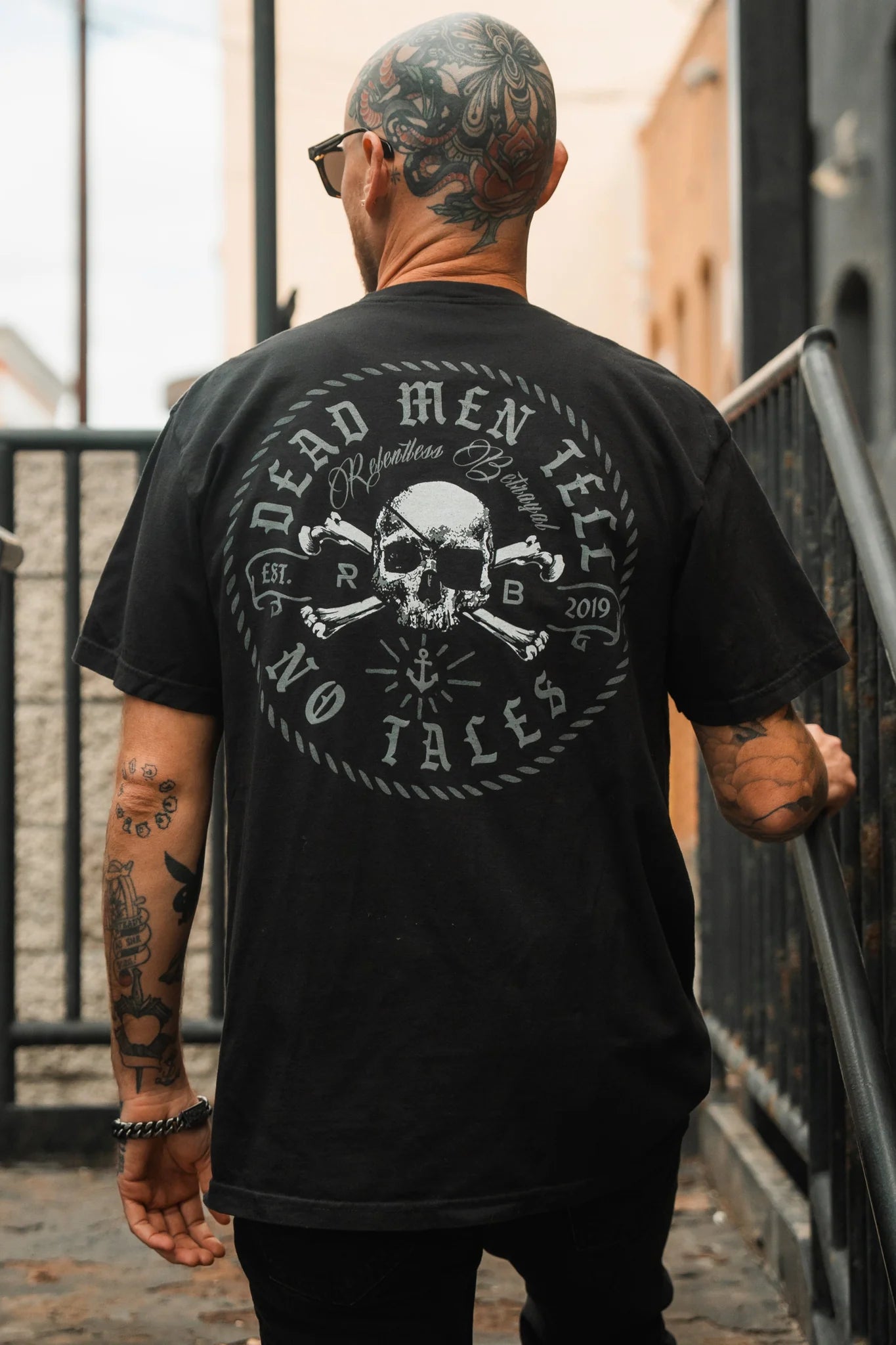 A bald man with tattoos is turned away from the camera, wearing the Relentless Betrayals Tell No Tales Premium Tee in black jersey cotton with a skull and crossbones design saying Dead Men Tell No Tales, standing near urban metal railings.