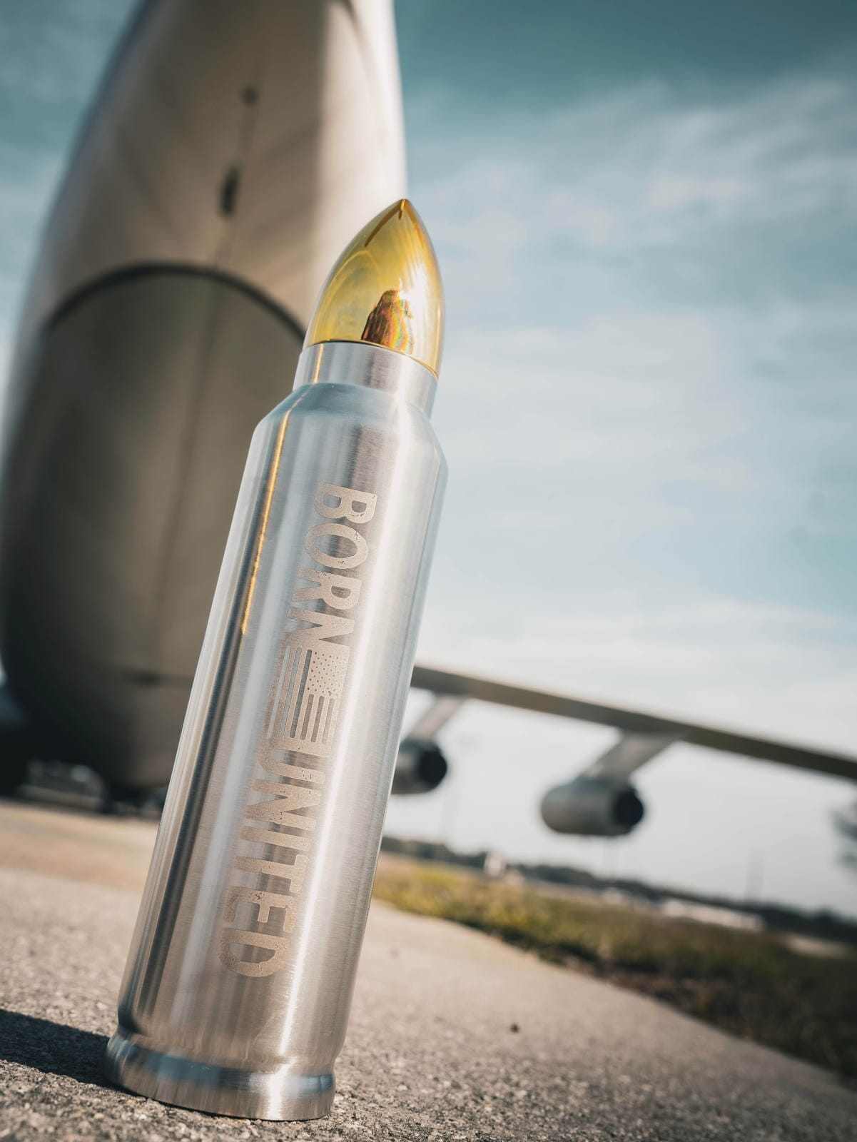 A large Bullet Thermos from Born United, featuring the BORN UNITED logo and an American flag design, sits in front of a massive airplane. Its presence under the clear blue sky embodies unity and freedom with every sip.