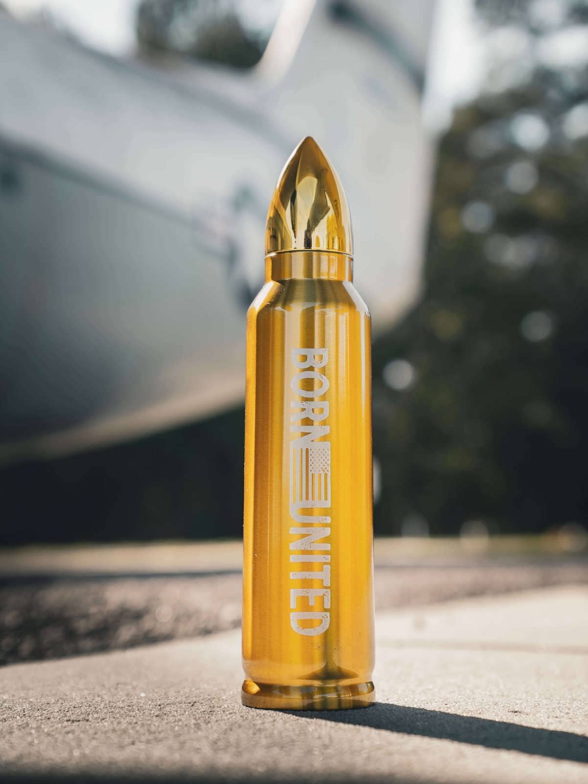 The Bullet Thermos by Born United Private Label, a gold bullet-shaped bottle with a tip-like top, stands out on the concrete. Blurred aircraft and trees in the background enhance its striking design, just like your first sip of Fives finest brew.