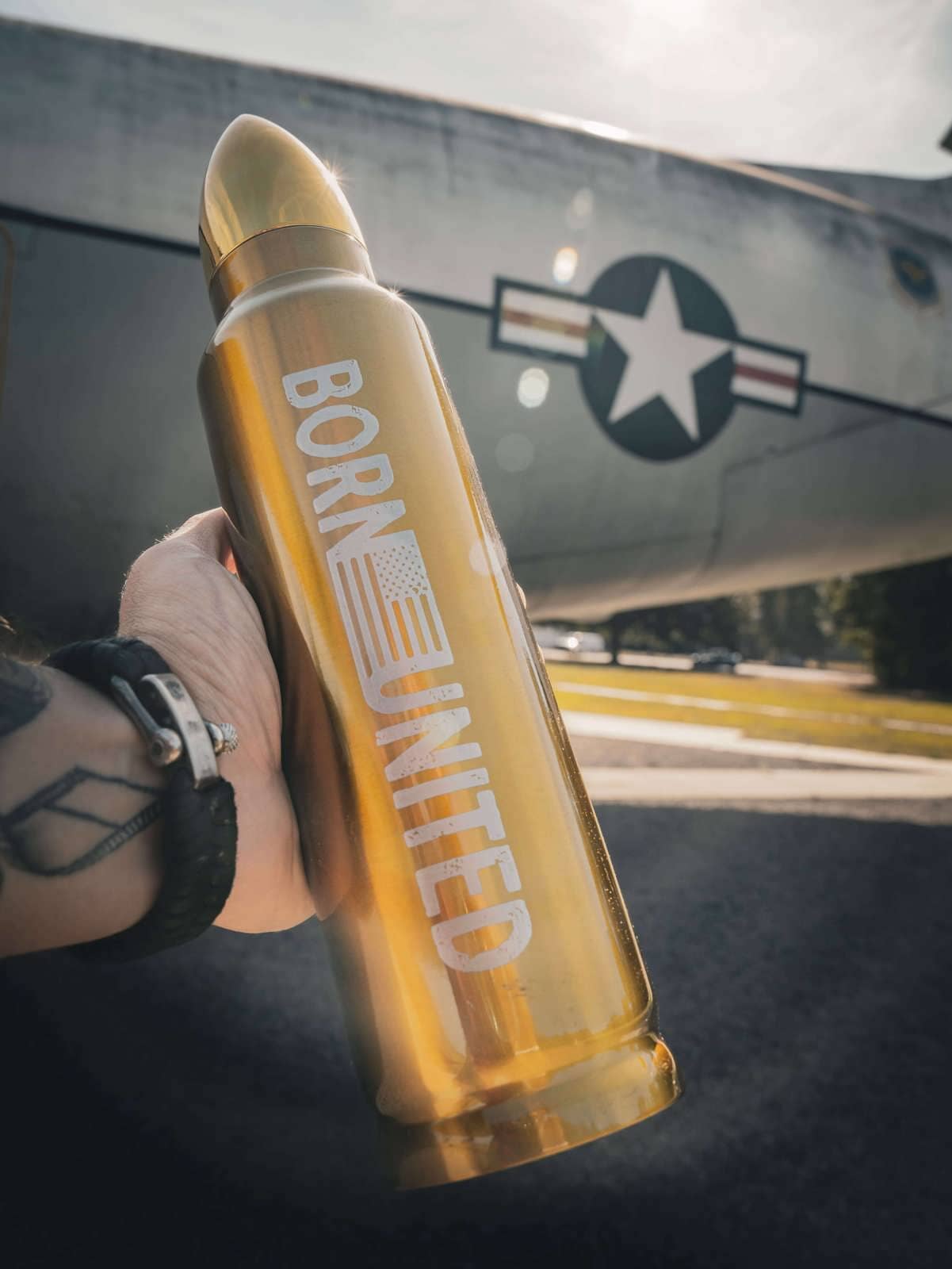 A person holds a bullet-shaped metallic Bullet Thermos by Born United Private Label with Five Sip on it, while in the background, an aircraft with a star insignia is visible outdoors on a sunny day.