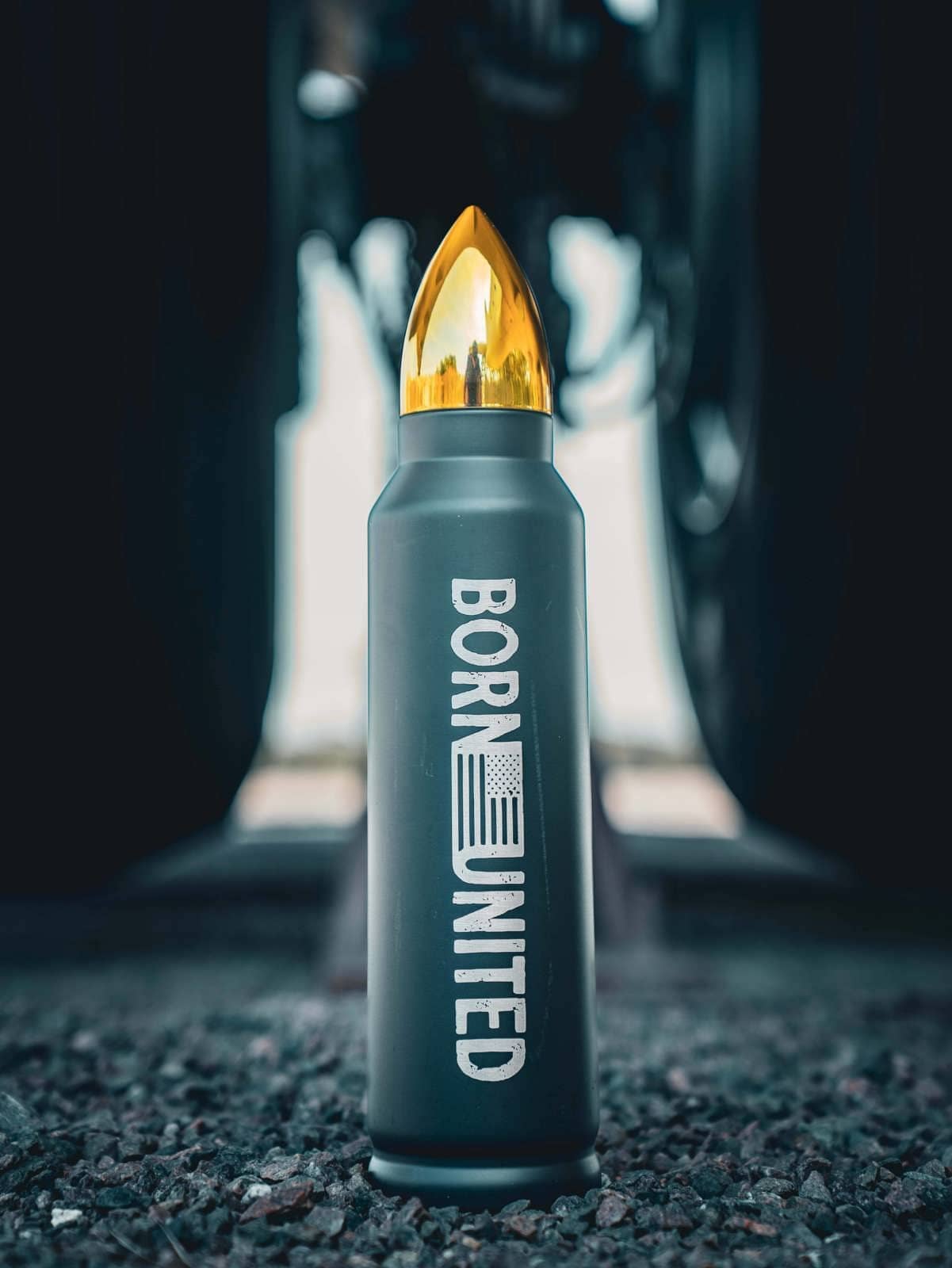 A Bullet Thermos by Born United Private Label rests on gravel, shaped like a bullet with Born United scripted along its side. Its cap resembles a golden bullet tip, set against a blurred industrial background to enhance its rugged appeal.