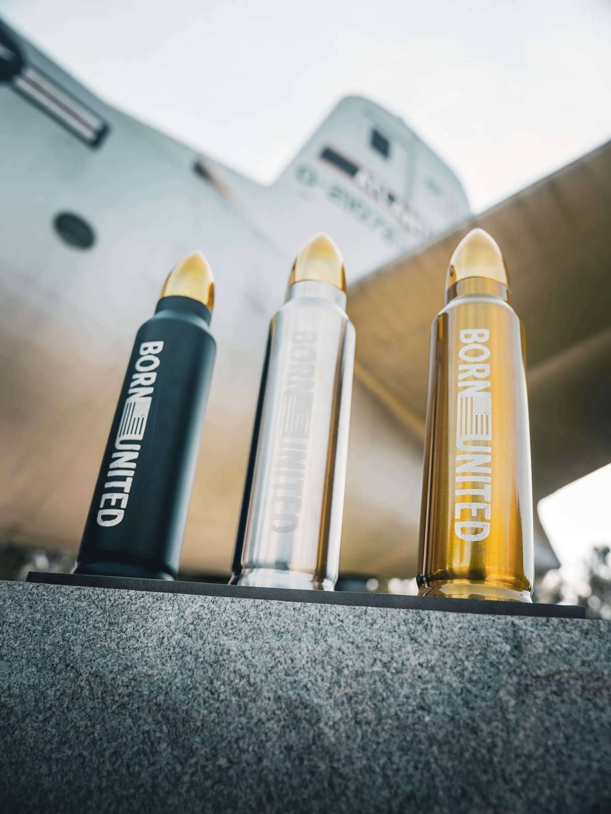 Three Bullet Thermos bottles by Born United Private Label rest on a stone surface in black, silver, and gold. A large aircraft looms behind with visible wings and fuselage. Each bottle promises a five-star experience in one sip.