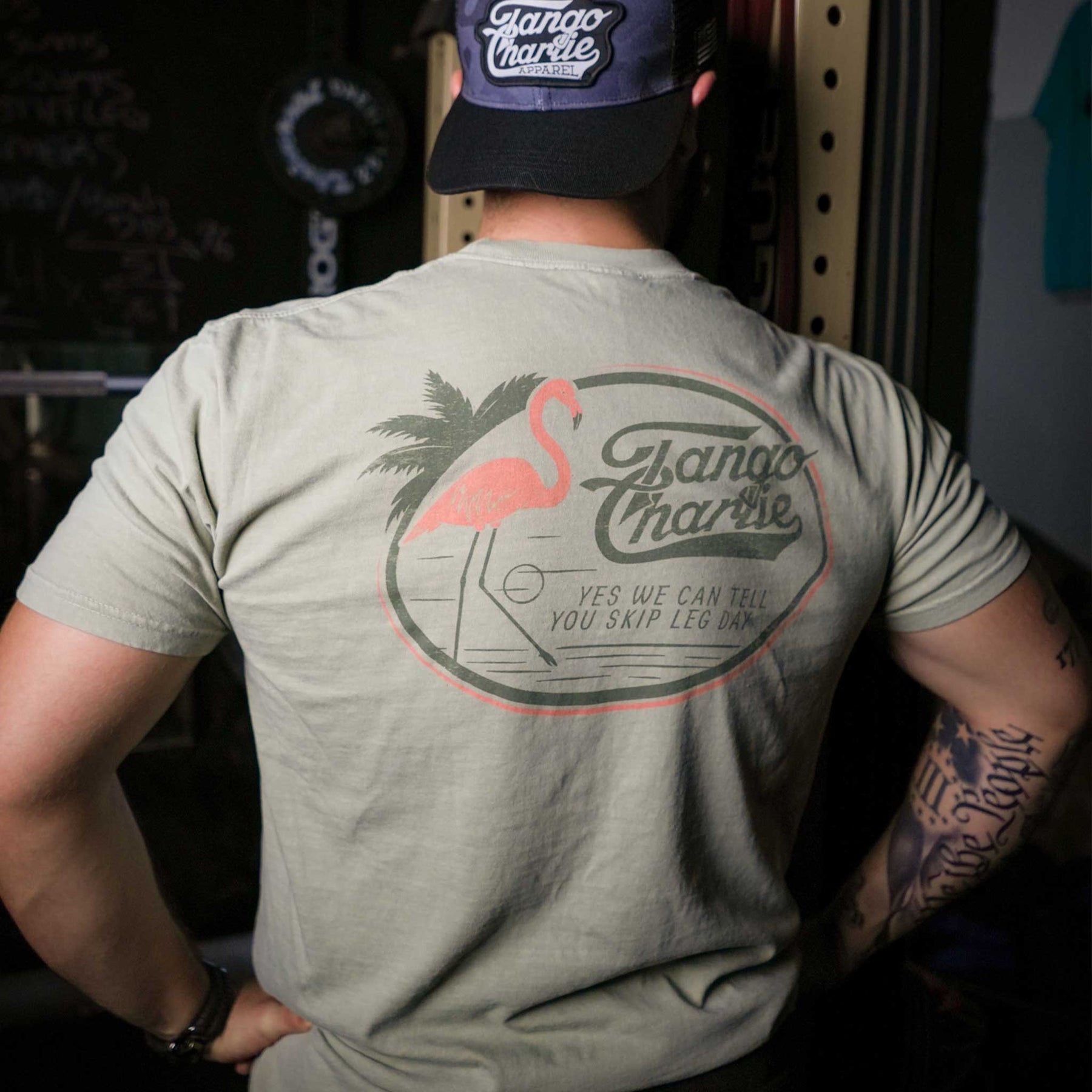 A person in a Tango Charlie Apparel Flamingo Legs Premium Tee and cap stands with their back to the camera. The beige shirt displays a pink flamingo, palm tree, and the text: YES WE CAN TELL YOU SKIP LEG DAY.