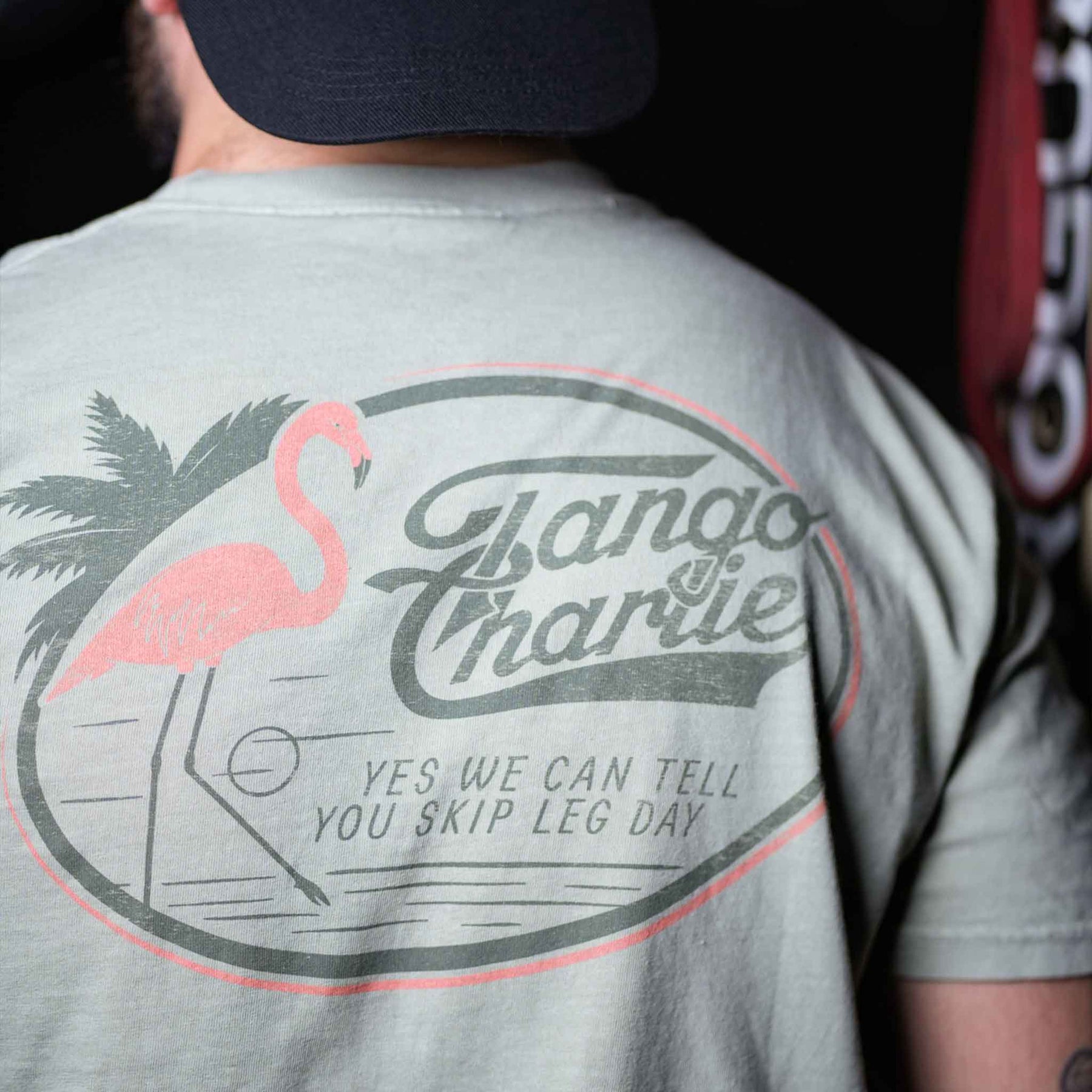 A person wearing the Flamingo Legs - Premium Tee by Tango Charlie Apparel, a breathable light-colored shirt with a flamingo, palm tree, and YES WE CAN TELL YOU SKIP LEG DAY printed on the back.