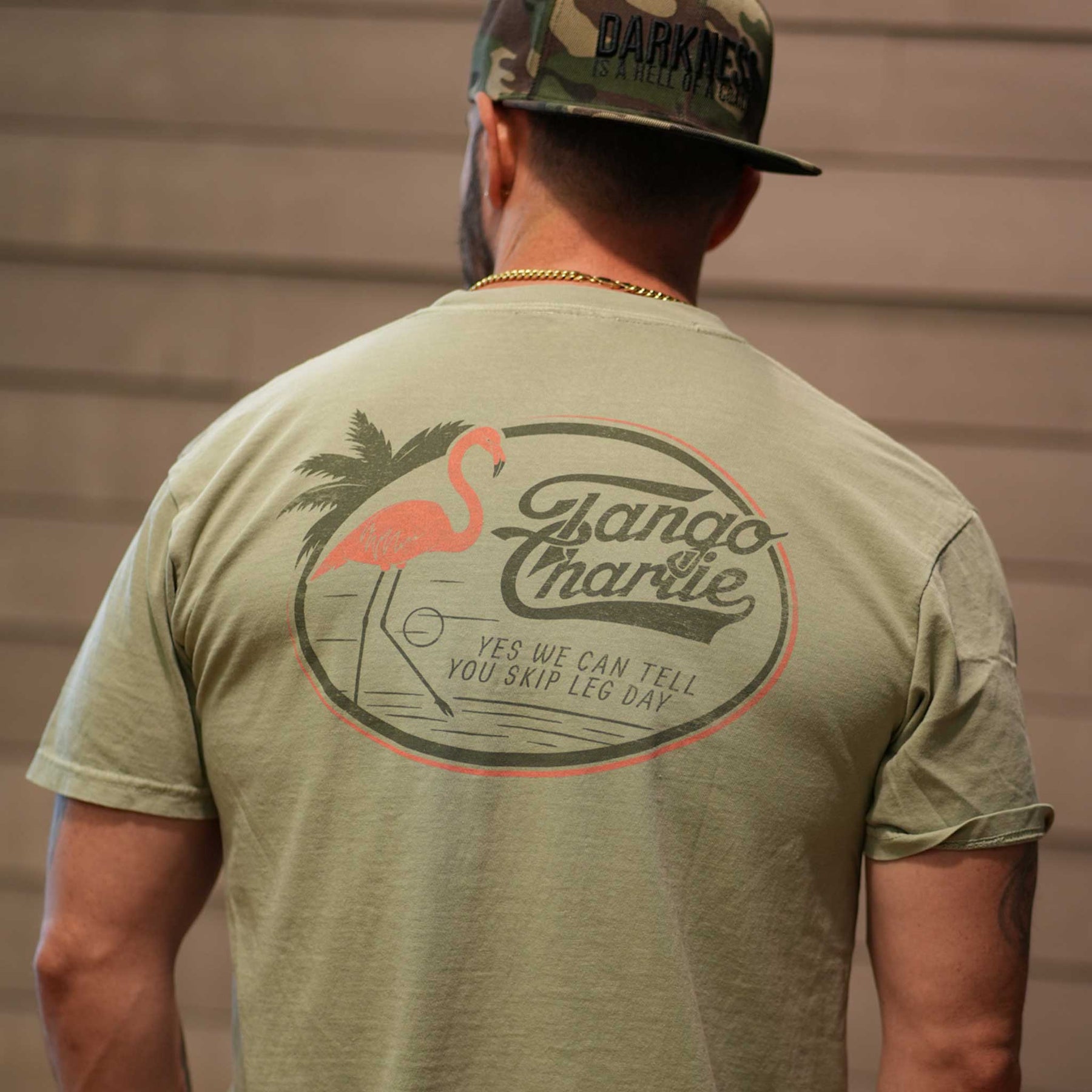 A man faces away from the camera in a Tango Charlie Apparel camo cap and their Flamingo Legs Premium Tee, featuring a flamingo and palm tree with Yes we can tell you skip leg day on the back. This breathable shirt blends comfort and style.