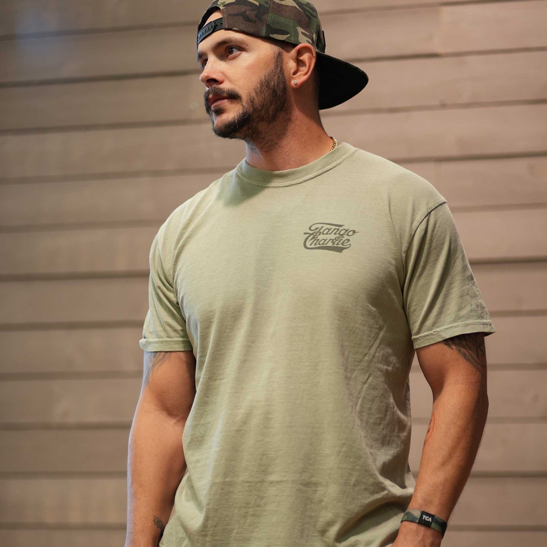 A bearded man wears a backward camouflage cap and a light green Flamingo Legs - Premium Tee by Tango Charlie Apparel, standing indoors against a wooden wall and looking to the side.