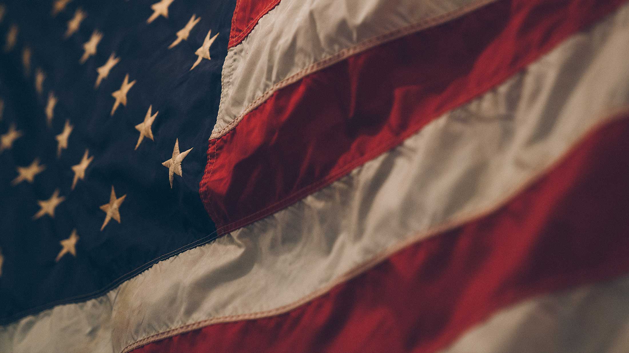 Close-up view of the United States flag showing the stars and stripes. The fabric appears slightly waving, displaying the red, white, and blue colors prominently. The focus is on the texture and patterns of the flag.