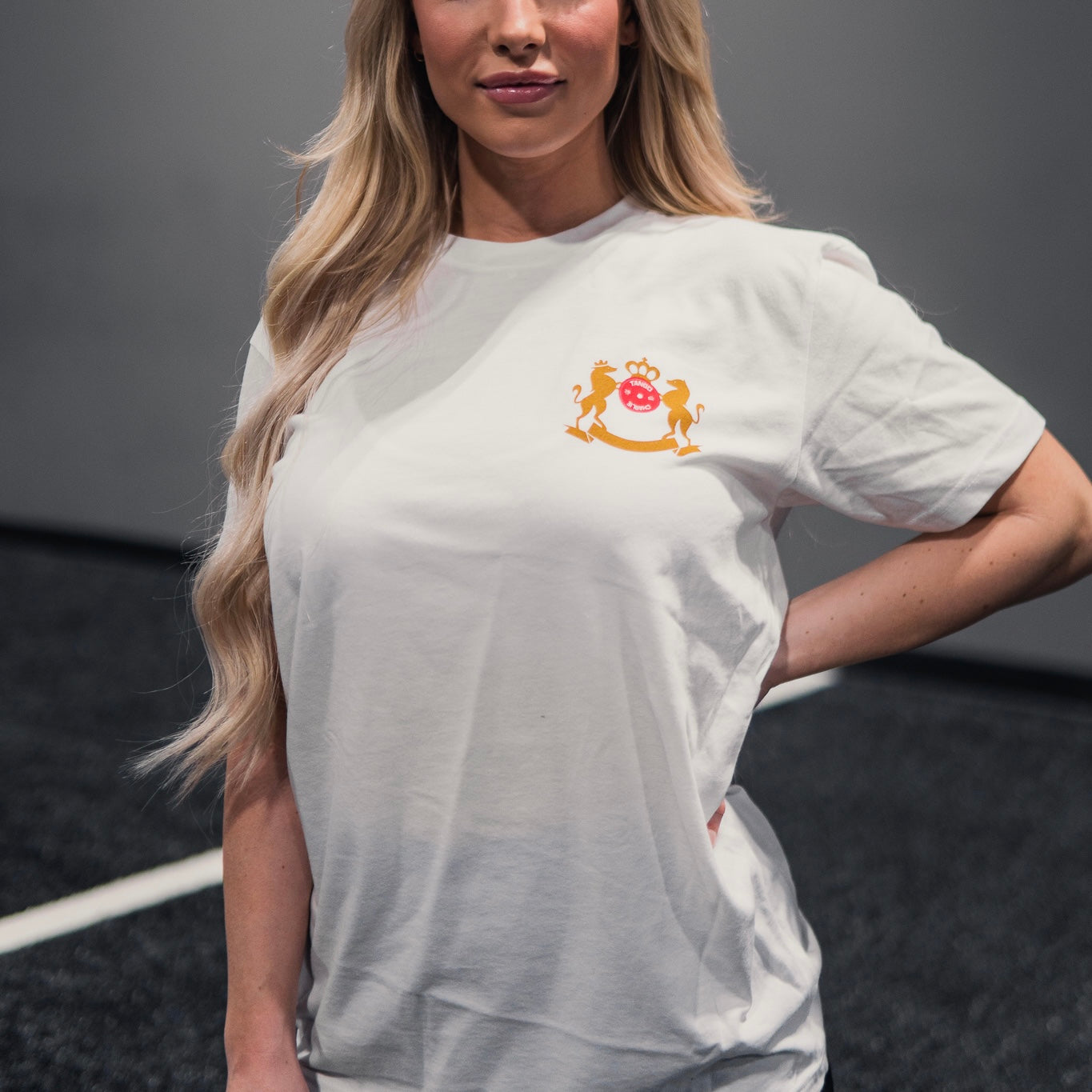 A woman with long blonde hair is wearing the Deadlift Addiction - Tee by Tango Charlie Apparel, featuring a colorful crest on the upper left. She stands indoors, hand on hip, against a plain backdrop.