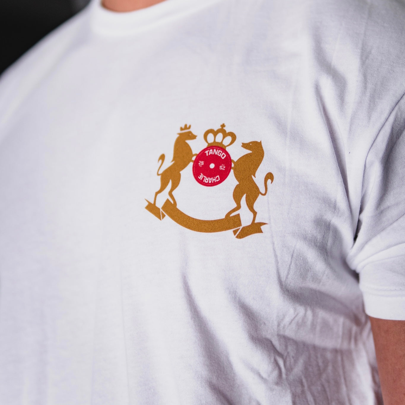 Close-up of a person in a Deadlift Addiction tee from Tango Charlie Apparel, featuring a gold crest with a lion and unicorn flanking a red circle that reads TANGO CHARLIE, alongside the lifter generals warning.