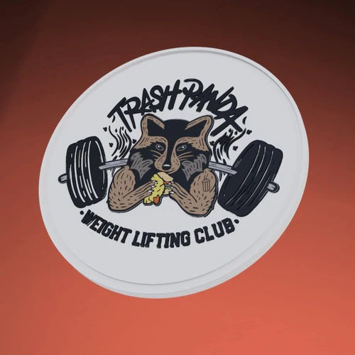 The Trash Panda Patch by Tango Charlie Apparel features a PVC design of a raccoon with a banana between dumbbells, set against a white circular background. TRASH PANDA is above and WEIGHT LIFTING CLUB below on an orange backdrop, with adhesive backing for easy application.
