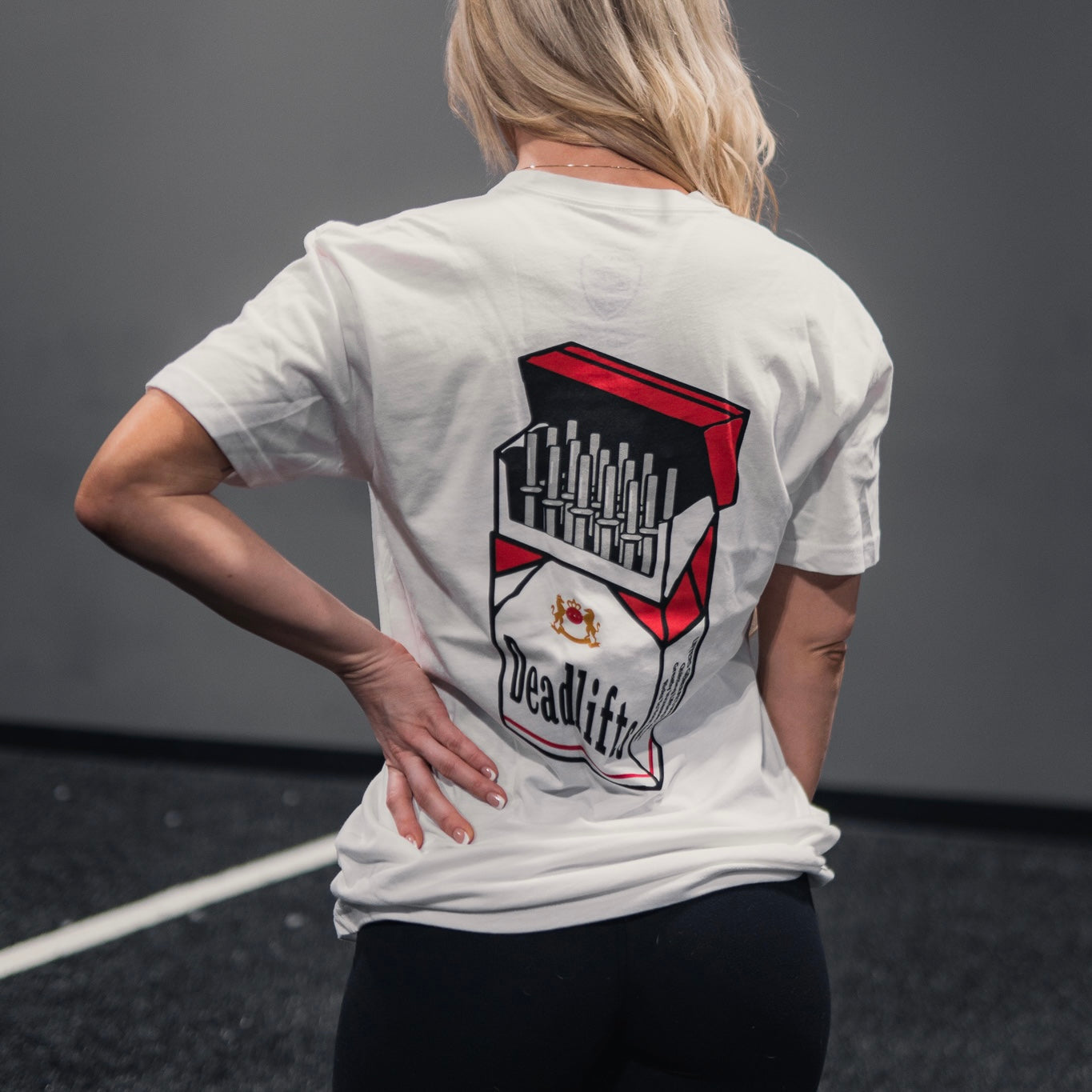 In a gym, a long-haired individual confidently wears the Deadlift Addiction - Tee by Tango Charlie Apparel, featuring the Deadlifts cigarette pack graphic on the back. With a hand on their hip, they embody the lifters warning: deadlifts can be seriously addictive.