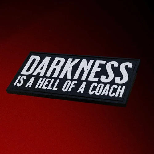 The Darkness Is A Hell Of A Coach Patch by Tango Charlie Apparel features white text on black against a dark red background. This striking PVC patch includes sewn hook backing for easy attachment to your favorite gear.