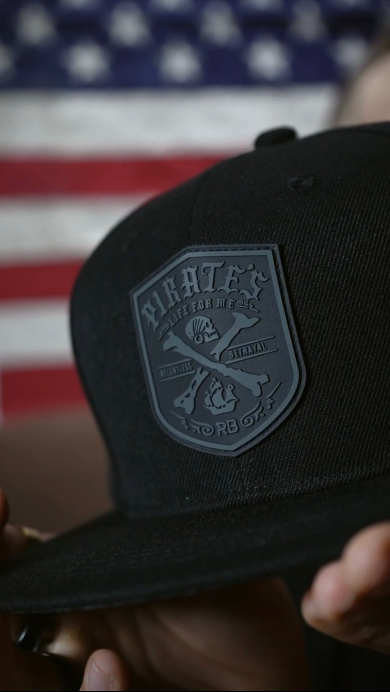 Close-up of the Pirates Life Black Snapback from Relentless Betrayal, featuring an embossed Rare shield logo with a fist and motor parts on a black cap with snapback closure, set against a blurred American flag backdrop.
