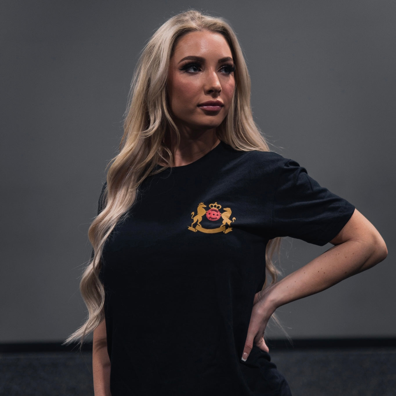 A person with long blonde hair confidently stands with one hand on their hip, dressed in Tango Charlie Apparels Deadlift Addiction Tee, featuring a crest adorned with a crown and lion emblem, against a plain background.