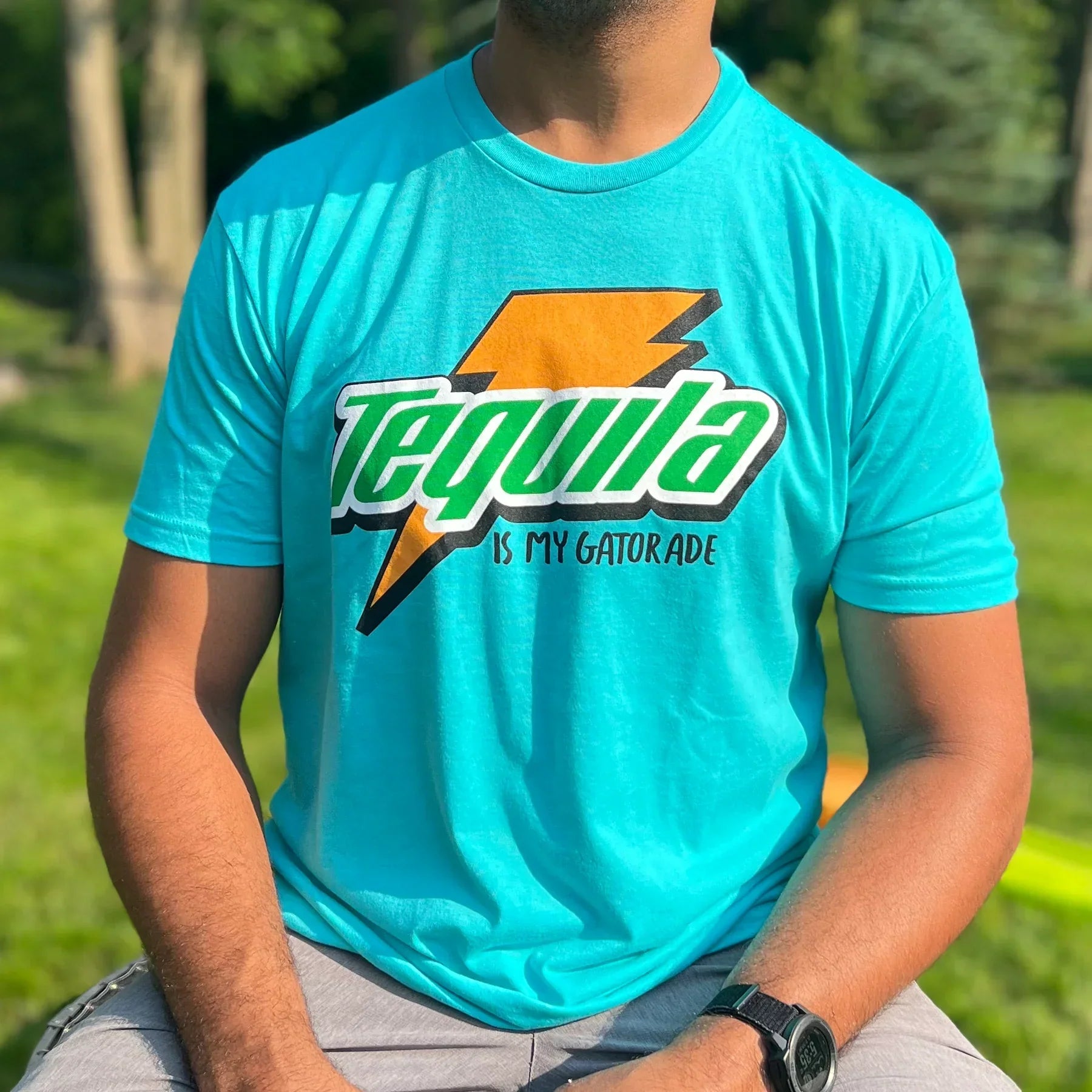 A person sits outdoors on a sunny day wearing the Tequila Is My Gatorade - Berry Blue T-shirt by Tango Charlie Apparel, featuring a bold yellow lightning bolt. The background shows vibrant green grass and trees.