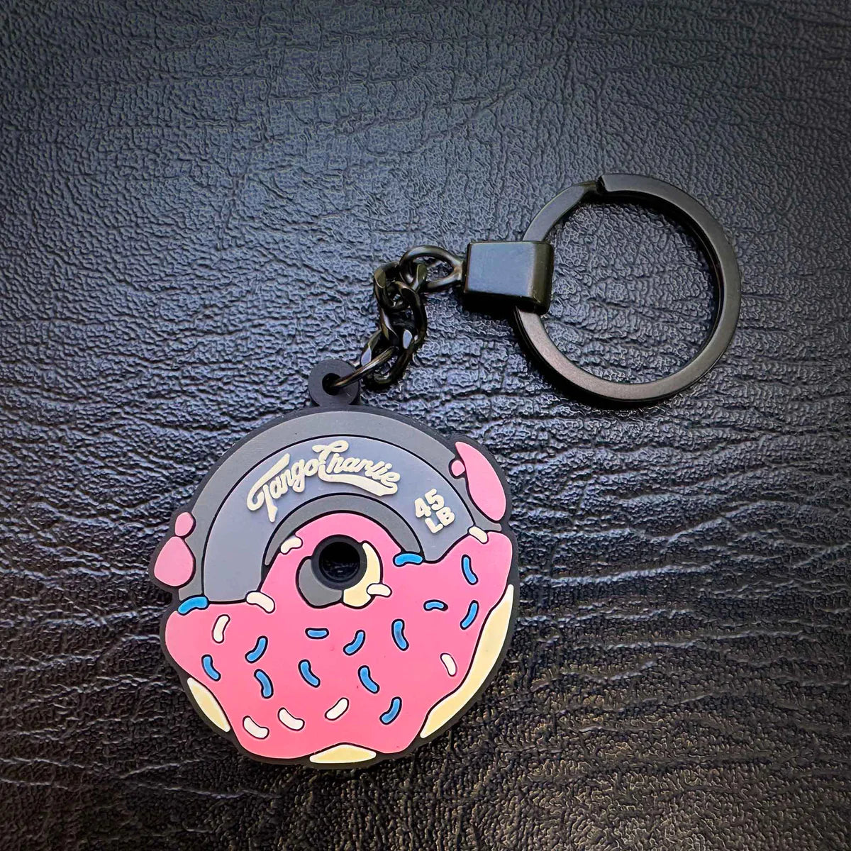A durable PVC keychain by Tango Charlie Apparel, the 45LB Plate Donut Keychain is shaped like a vinyl record with Tango Charlie printed on it. It features a rubber design resembling a donut with pink frosting and blue and white sprinkles, all set against a textured black background.