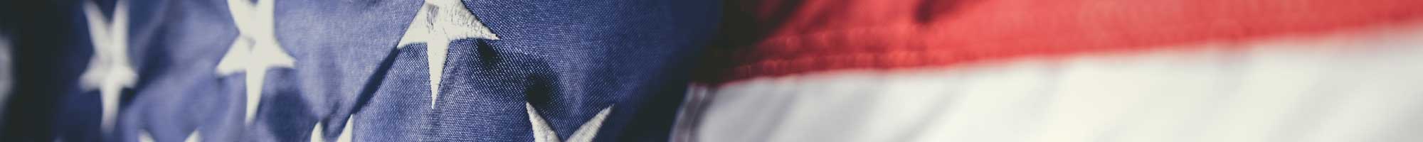 Close-up of a weathered American flag showing the blue field with white stars on the left and red and white stripes on the right. The texture suggests an aged or vintage appearance.