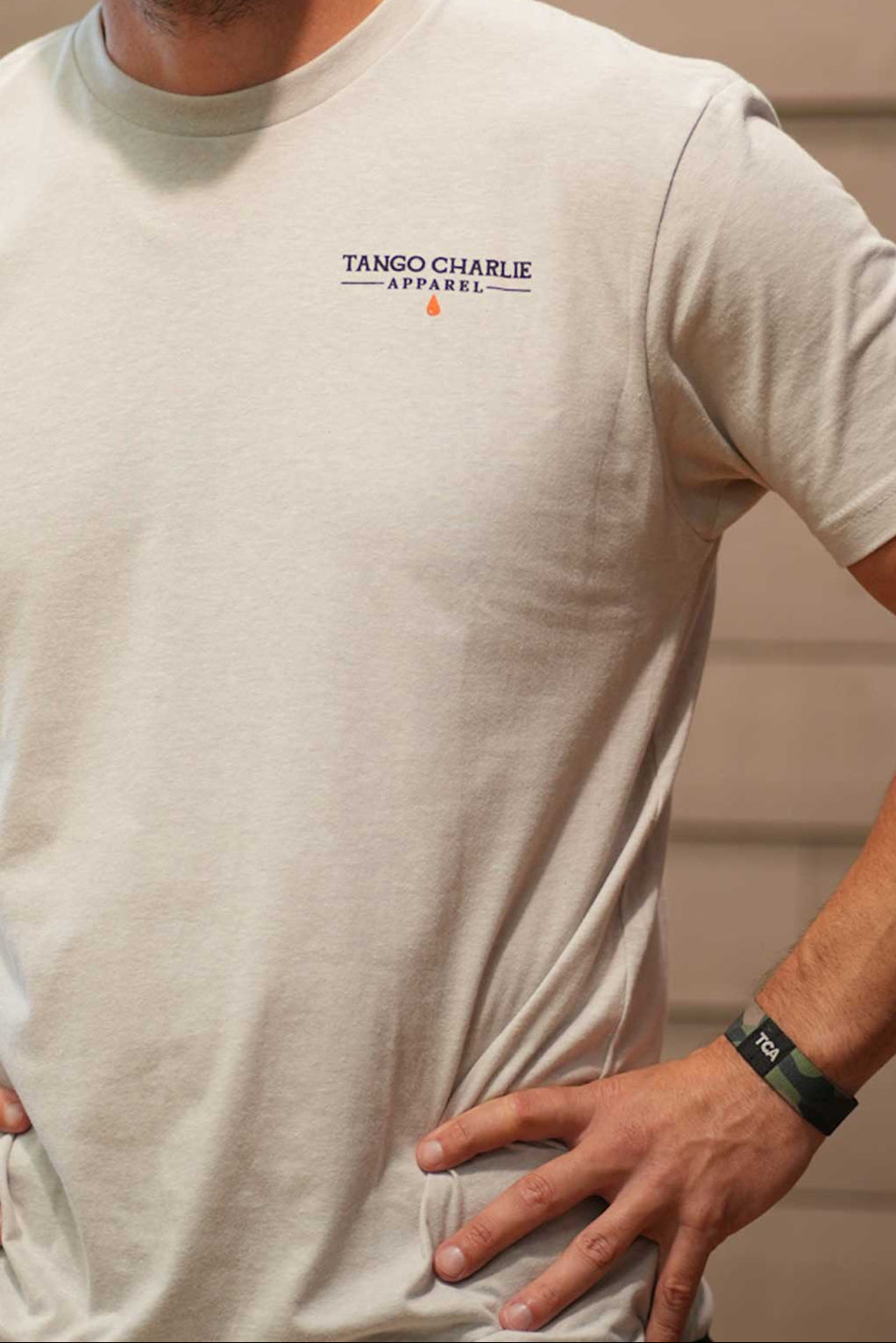 A bearded man in a backward cap stands hands on hips before a wooden wall, wearing the light-colored Time's Up Tee by Tango Charlie Apparel, made from ring-spun cotton and featuring a motivational design.