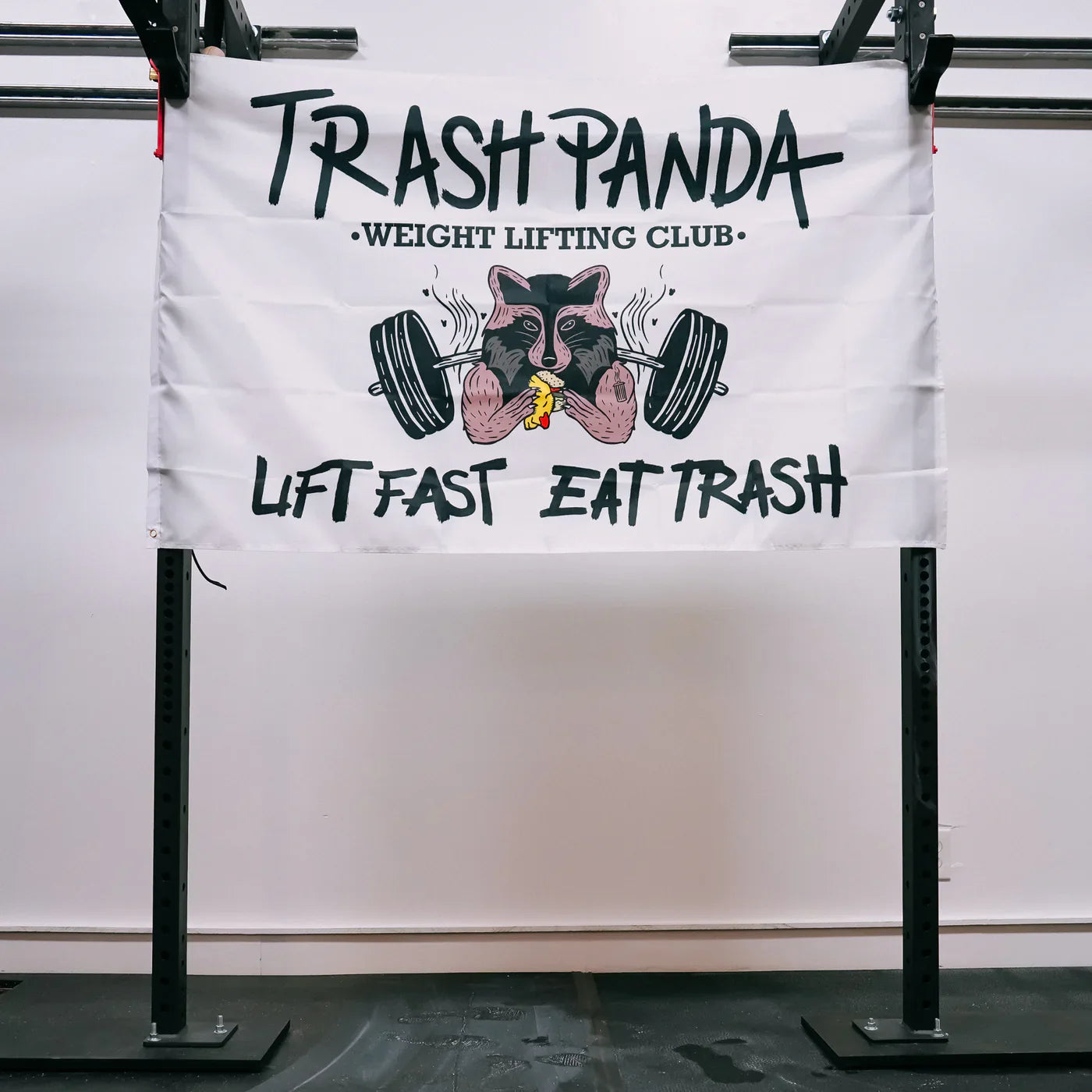 A polyester flag from Tango Charlie Apparel hangs on the squat rack, featuring Trash Panda Weight Lifting Club, a muscular cartoon raccoon lifting weights with a banana, and the bold slogan Lift Fast, Eat Trash.