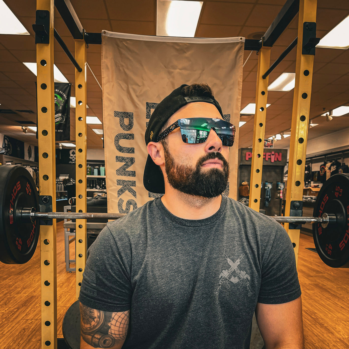 A bearded man in a grey t-shirt and backward cap sports Drippy Black Sunglasses by Drippy Swimwear, sitting before a barbell at the gym with a banner and equipment in the background.