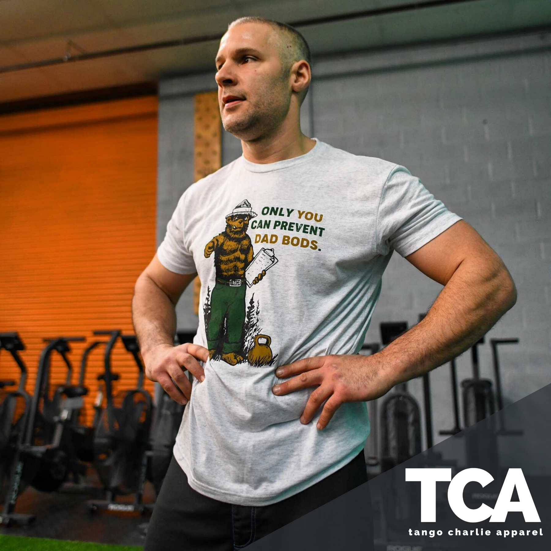 A person stands confidently in a gym wearing the Only You Can Prevent Dad Bod T-Shirt by Tango Charlie Apparel featuring a cartoon bear. Exercise equipment fills the background, with Tango Charlie Apparel and their logo displayed at the bottom right.