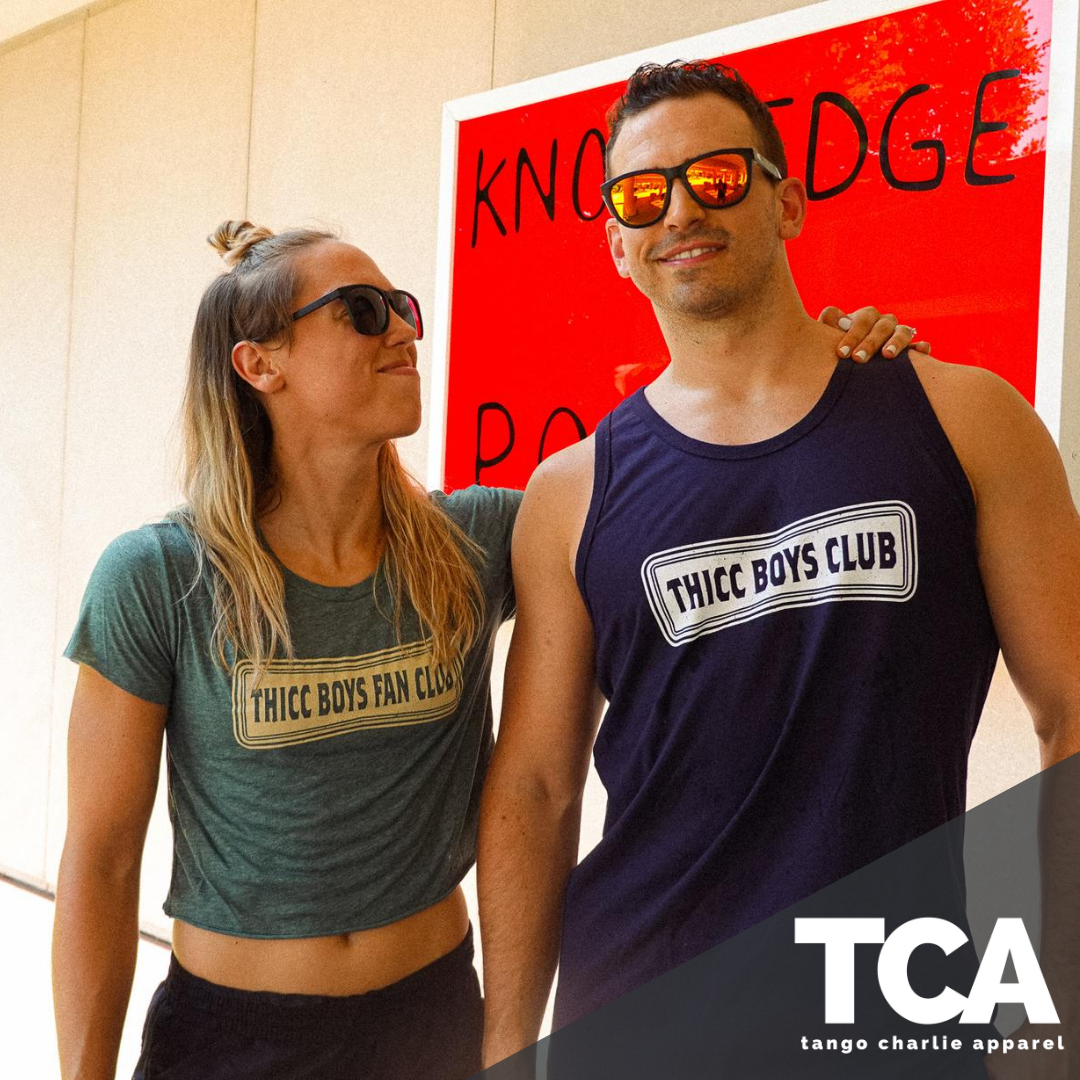 Two people wear Tango Charlie Apparels THICC Boys Fan Club Crop T-Shirts, along with stylish sunglasses as they smile in front of a red and white sign. The TCA logo is in the corner.