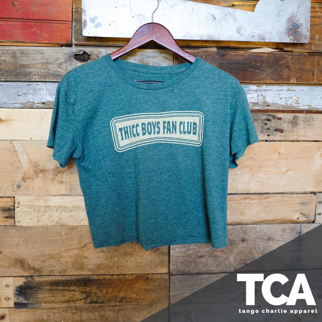 A green THICC Boys Fan Club Crop T-Shirt by Tango Charlie Apparel hangs stylishly on a wooden wall with the brands logos TCA and Tango Charlie Apparel in the bottom right corner.