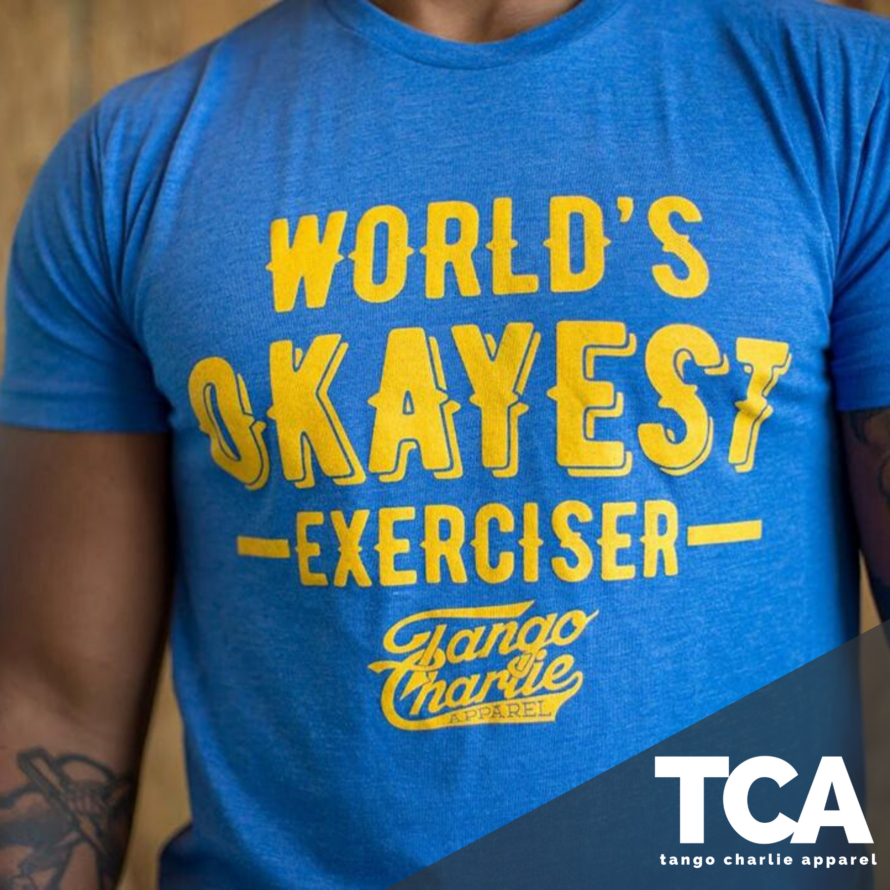 A person wears the comfy Worlds OKAYEST Exerciser Tee in blue, featuring yellow text and the Tango Charlie Apparel logo, showcasing a relaxed style.