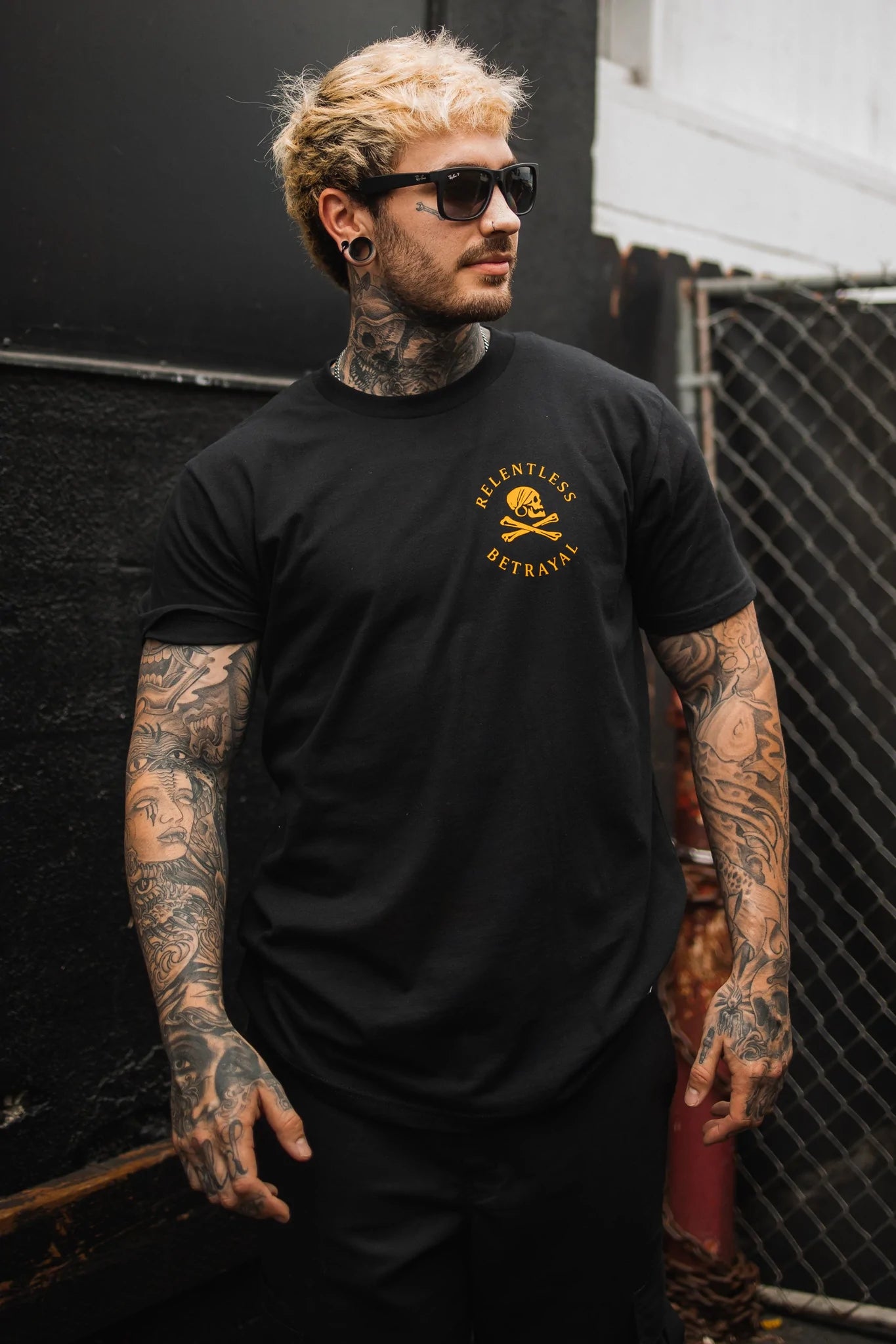 A tattooed man in sunglasses stands against a chain-link fence wearing a sleek Relentless Betrayal Lost At Sea Black T-shirt made from fine jersey cotton. The black tee with a yellow design on the left chest highlights his inked arms and neck, all while he maintains a serious expression.