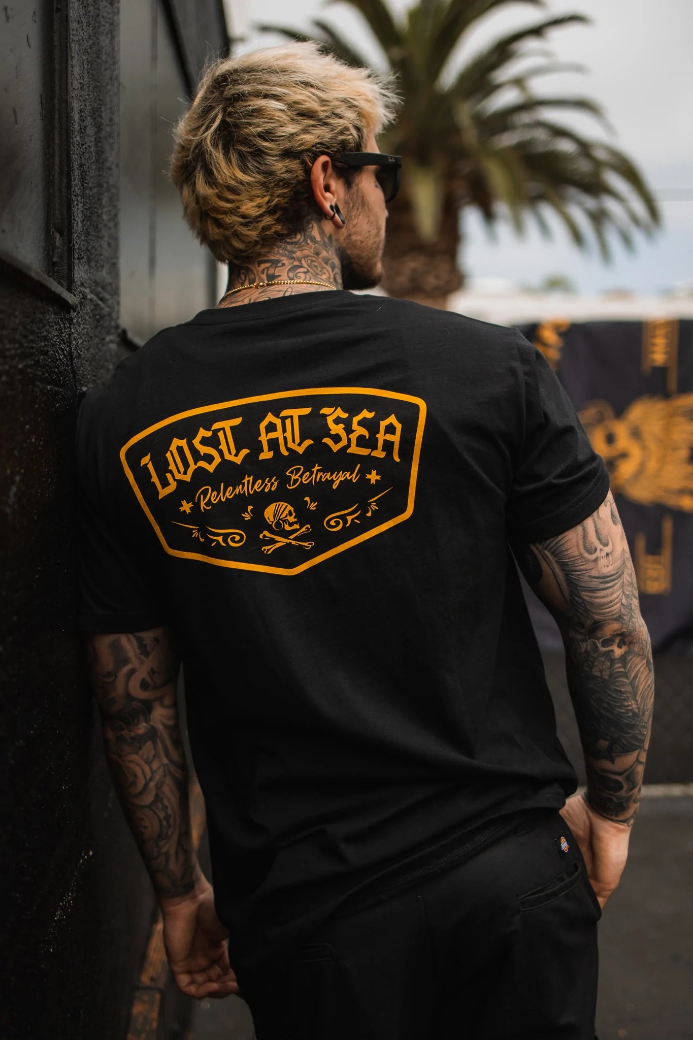 A tattooed individual with light hair is outdoors wearing sunglasses and a black Relentless Betrayal Lost At Sea UNISEX T-shirt. The fine jersey cotton tee features the phrase in orange on the back. Nearby, palm trees sway with a blurred flag in the distance.