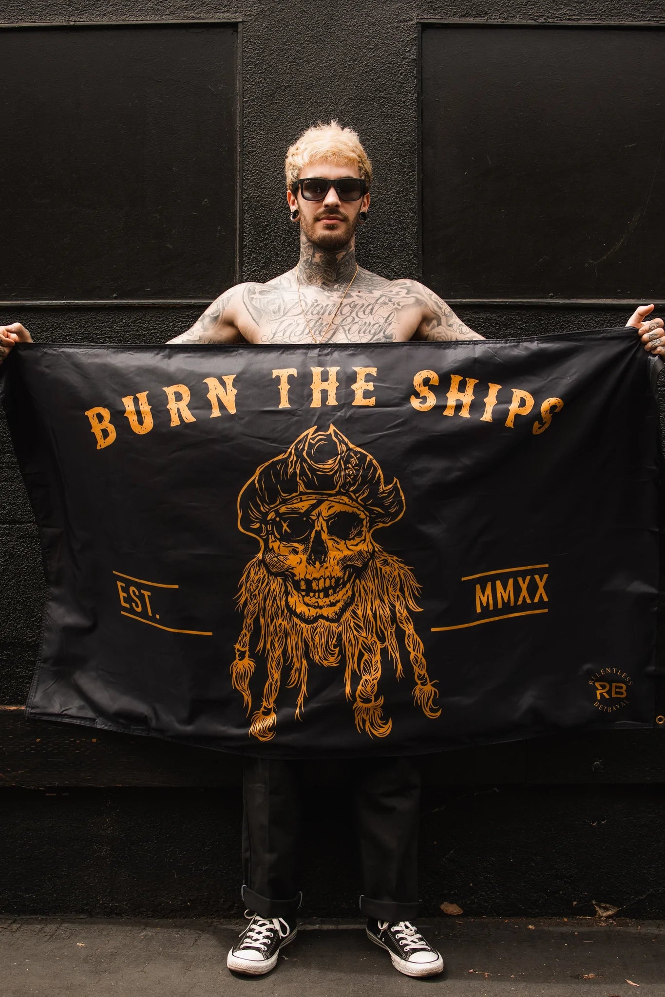 A tattooed individual with sunglasses holds Relentless Betrayals Burn The Ships BLACKOUT Double Sided Flag, crafted from durable polyester, featuring a yellow pirate skull and crossbones on a black background with BURN THE SHIPS and EST. MMXX, exuding classic pirate flair.