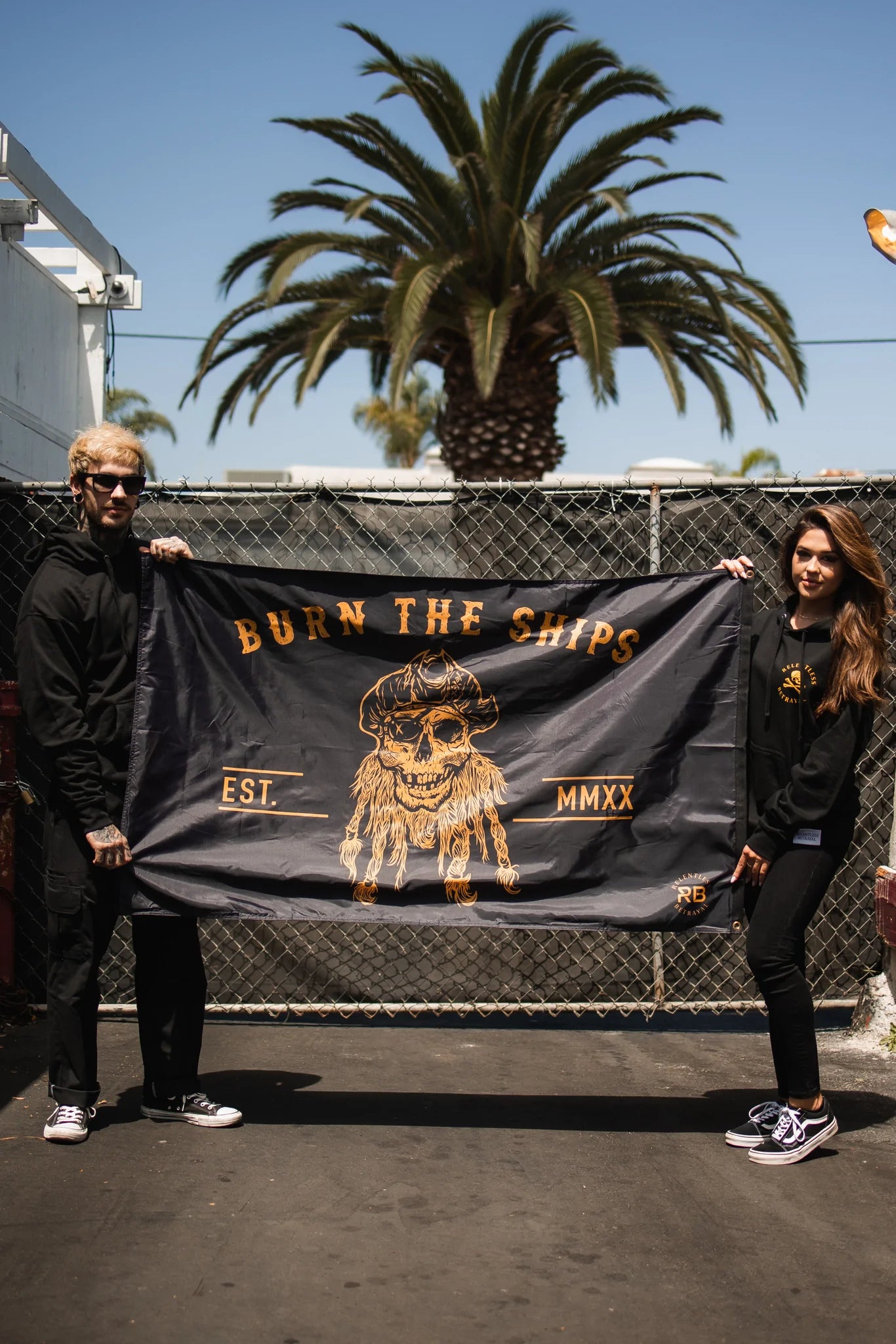 Two people hold a Relentless Betrayal Burn The Ships BLACKOUT Double Sided Flag, made from durable polyester with a stylish pirate skull and EST MMXX text, against a chain-link fence. A palm tree sways under the blue sky as both individuals, in black and sunglasses, stand confidently.