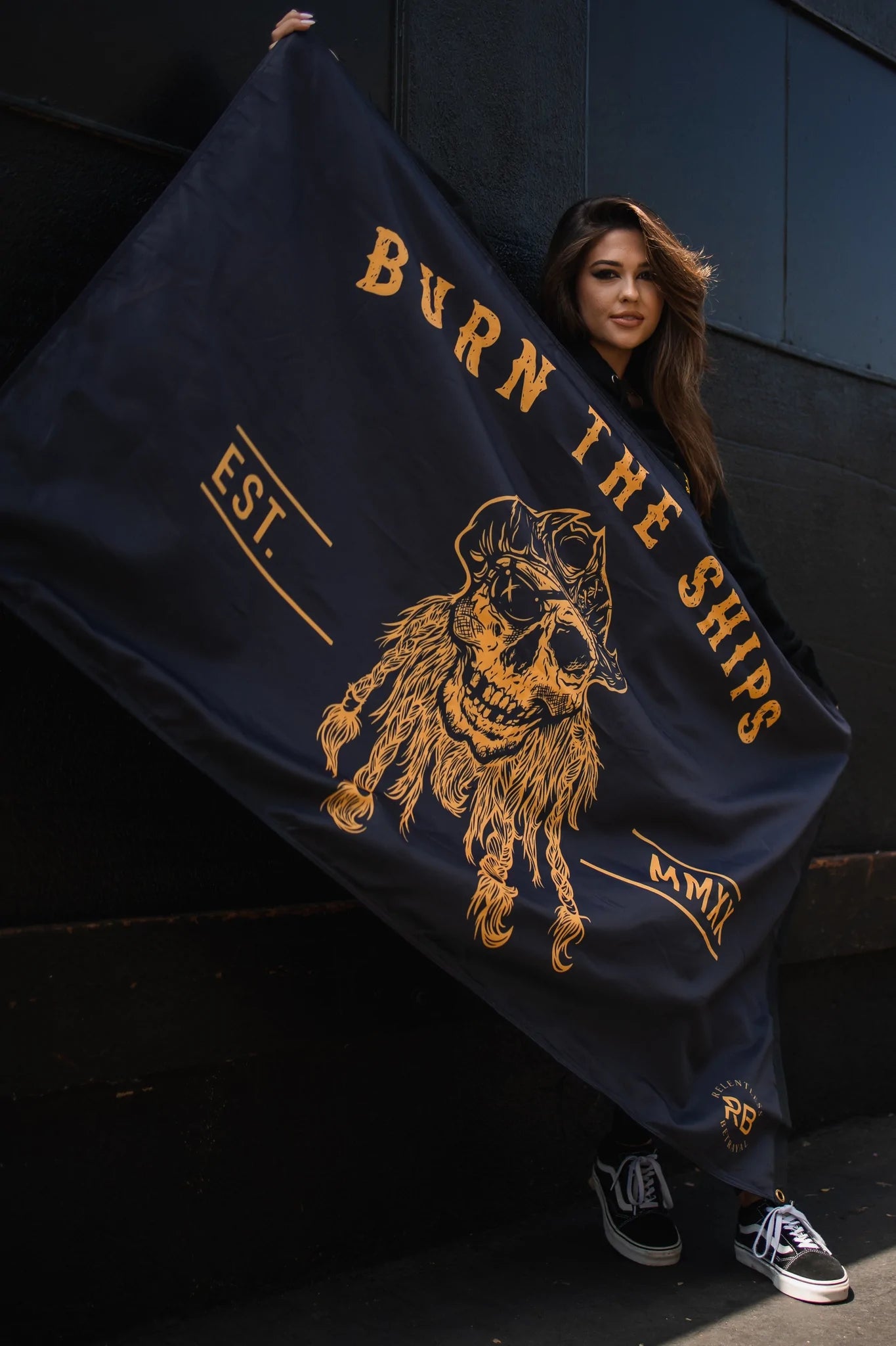 Someone holds the Burn The Ships BLACKOUT Double Sided Flag by Relentless Betrayal, made from premium polyester. It features a bearded pirate skull in gold with Burn the Ships, EST., and MMXIV. They stand casually against a dark wall.