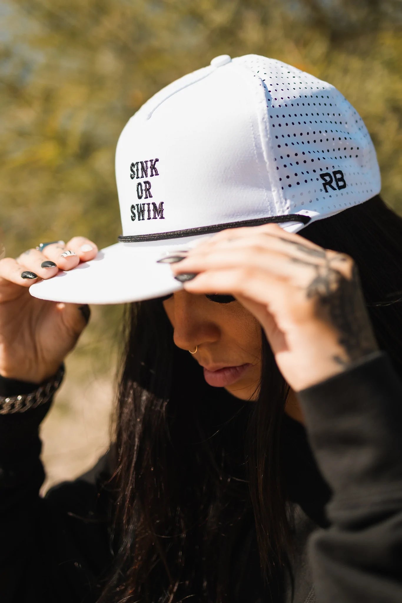 They adjust their Sink or Swim WHITEOUT Snapback by Relentless Betrayal, a white breathable cap featuring SINK OR SWIM and RB on the side, while wearing a black long-sleeve top against a backdrop of blurred greenery.