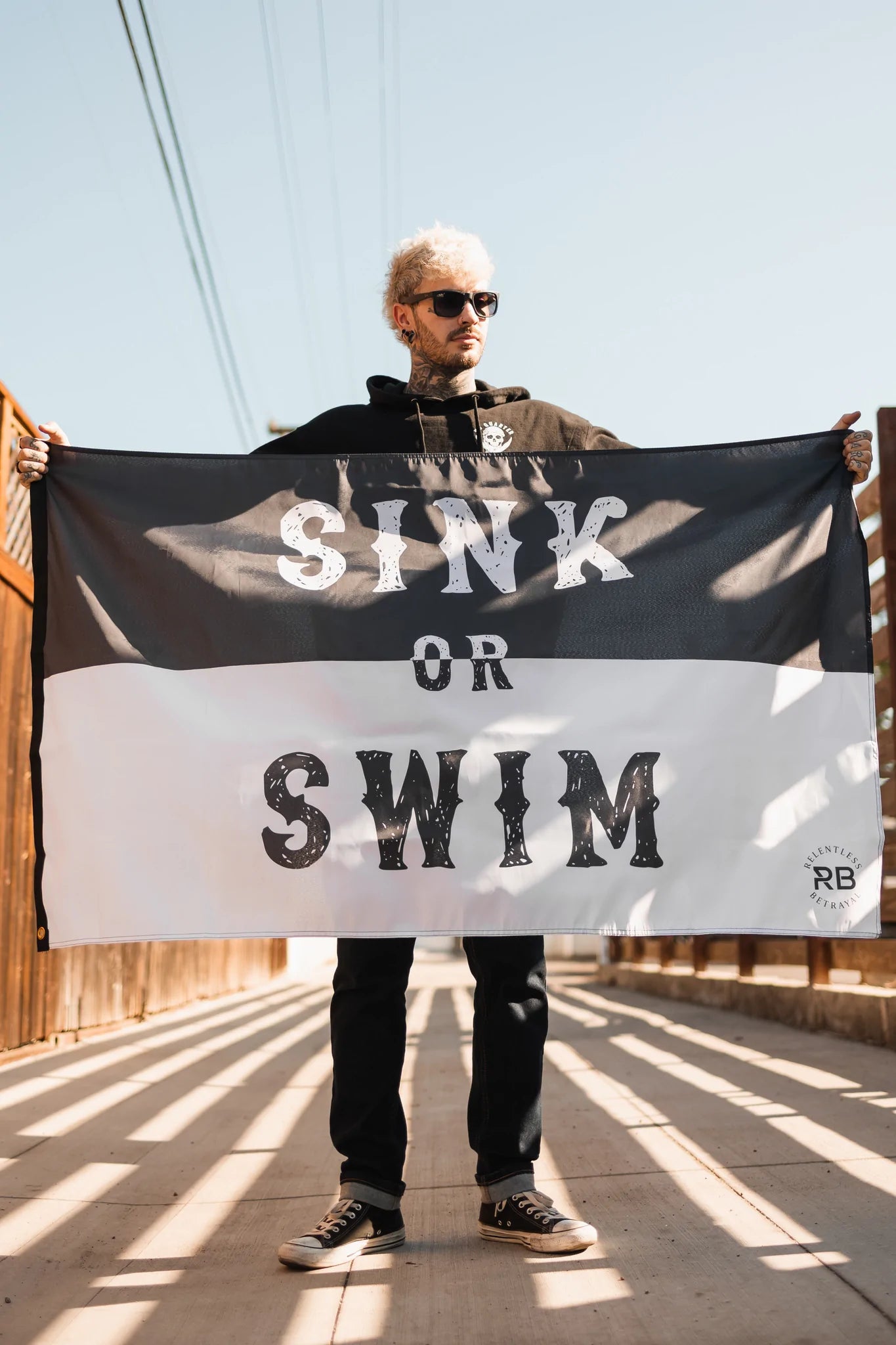 A person with sunglasses and light hair holds a Relentless Betrayal Sink or Swim Flag made of durable polyester on a sunlit wooden bridge, wearing a dark hoodie and jeans. Metal grommets glint under the clear blue sky.