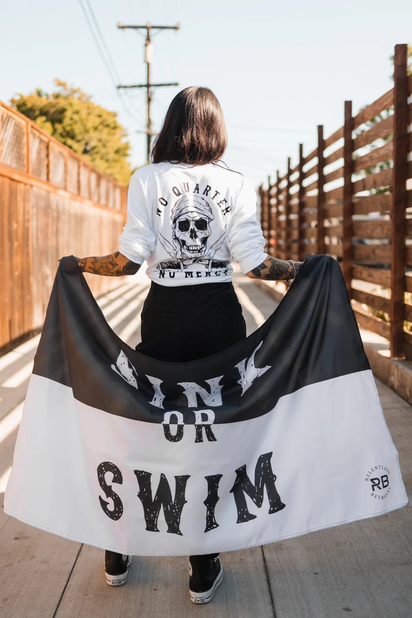 A tattooed person stands on a bridge, raising the Relentless Betrayal Sink or Swim flag made of durable polyester with metal grommets. They wear a long-sleeved skull-design shirt with text on the back.