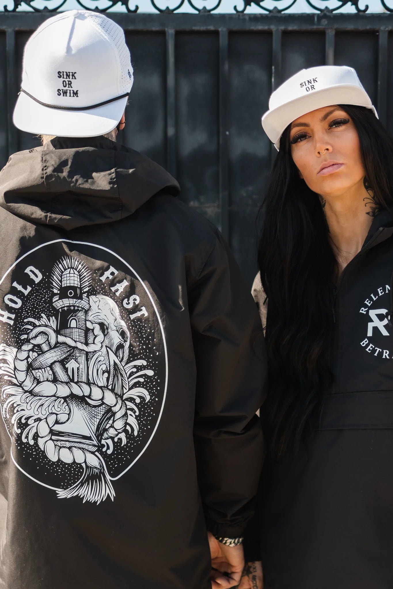 Two people stand side by side, wearing black jackets with white designs and sleek, breathable snapbacks. One jackets back shows a nautical graphic with HOLD FAST. Their Relentless Betrayal caps read SINK OR SWIM and SINK OR, named Sink or Swim WHITEOUT Snapback.