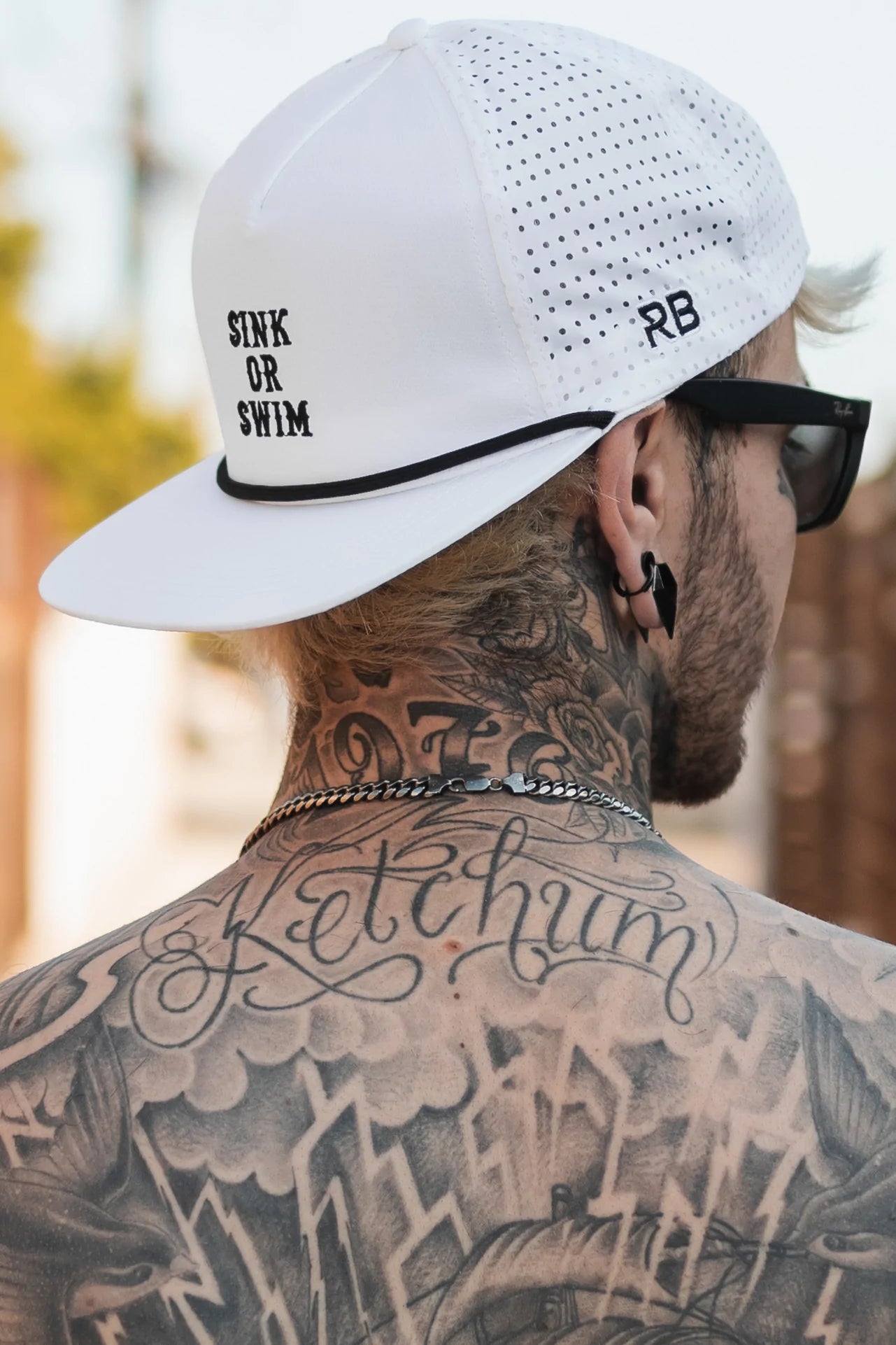 A tattooed and pierced individual is seen from behind wearing the Relentless Betrayals Sink or Swim WHITEOUT Snapback, paired with sunglasses and a chain necklace, showcasing detailed upper back tattoos.