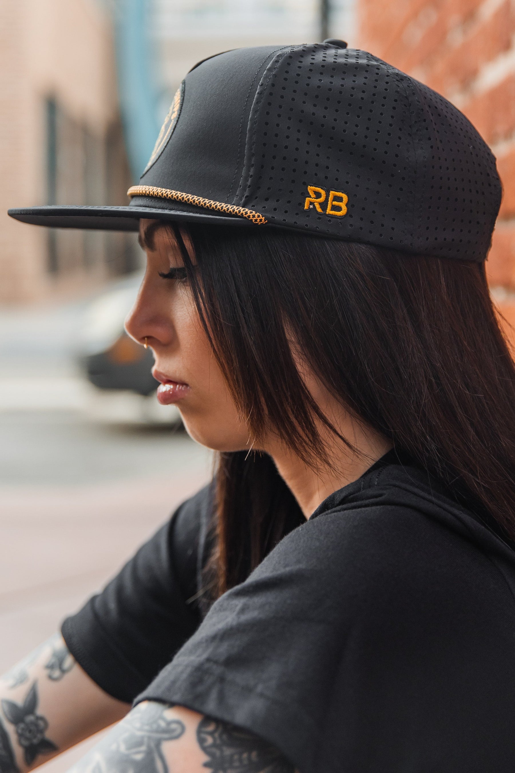 Against a brick wall, a person with long dark hair and tattoos sits contemplatively. They wear the Relentless Betrayal Burn The Ships BLACKOUT Snapback, featuring RB embroidered in yellow. An out-of-focus street scene forms the backdrop.