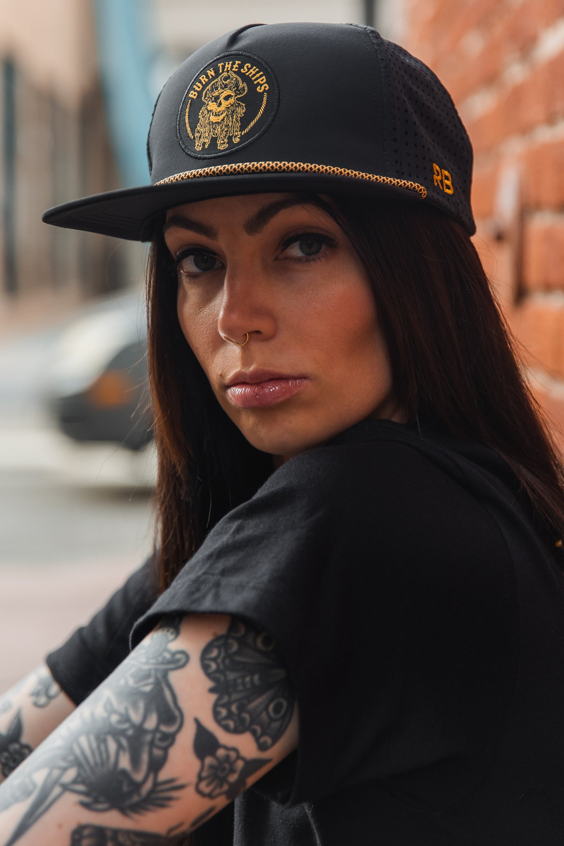 A person with long dark hair, tattoos on their arm, and a nose piercing is wearing the Burn The Ships BLACKOUT Snapback by Relentless Betrayal with gold skull embroidery. They are dressed in a black shirt and sit beside a brick wall.