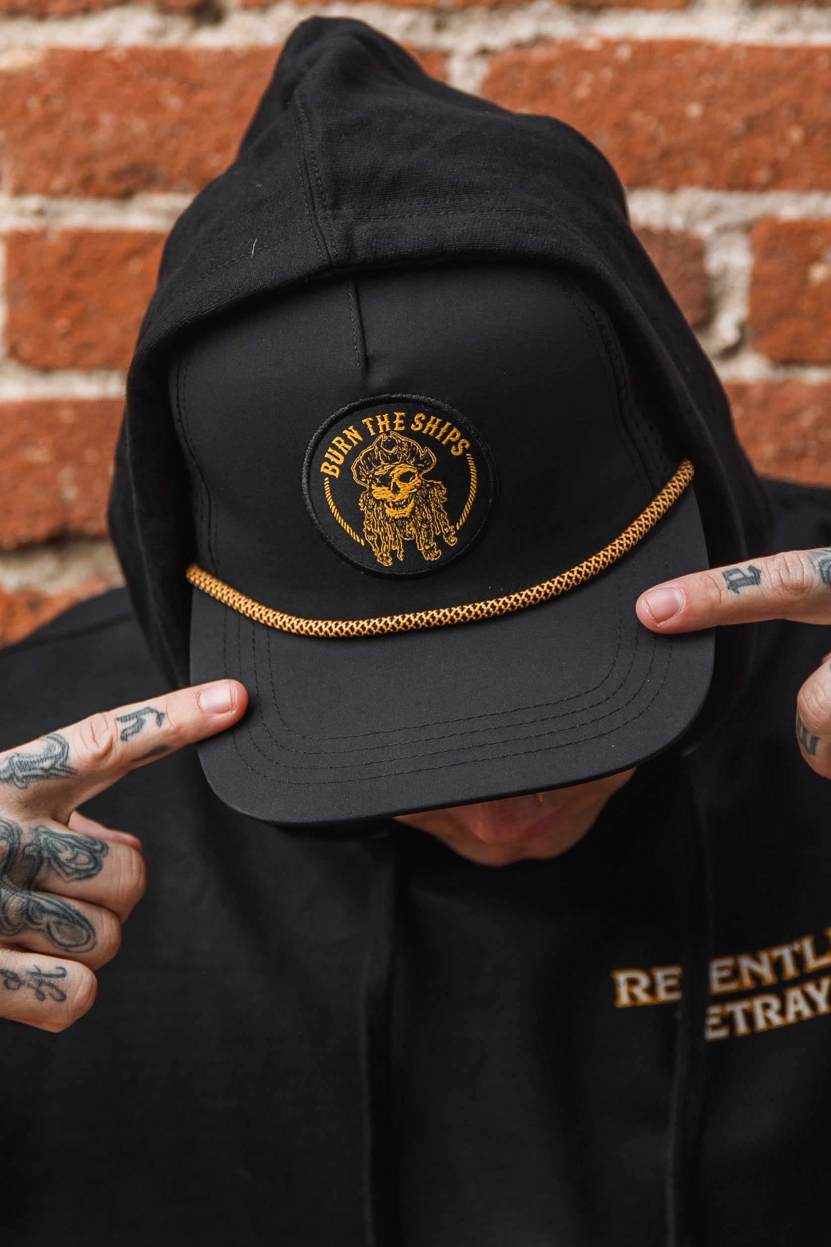 Wearing Relentless Betrayal’s Burn The Ships BLACKOUT Snapback, featuring a gold emblem, a tattooed individual adds edgy flair by posing against a brick wall and pointing at the cap.