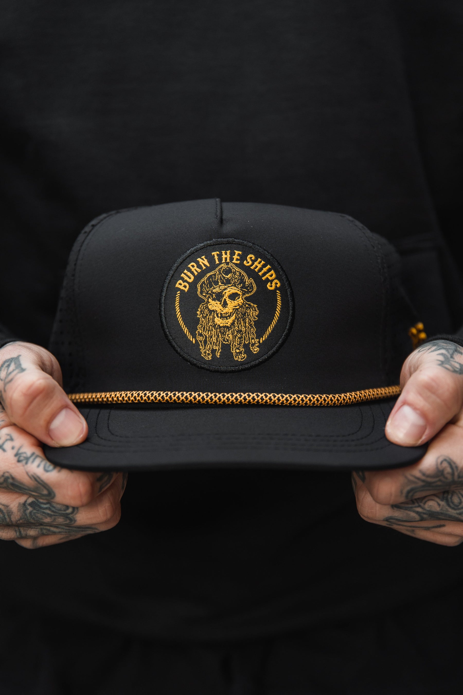 A person with tattooed hands holds the Relentless Betrayals Burn The Ships BLACKOUT Snapback, featuring a circular patch of a skull in a pirate hat with Burn the Ships in gold. A decorative gold chain above the brim completes this unique design.
