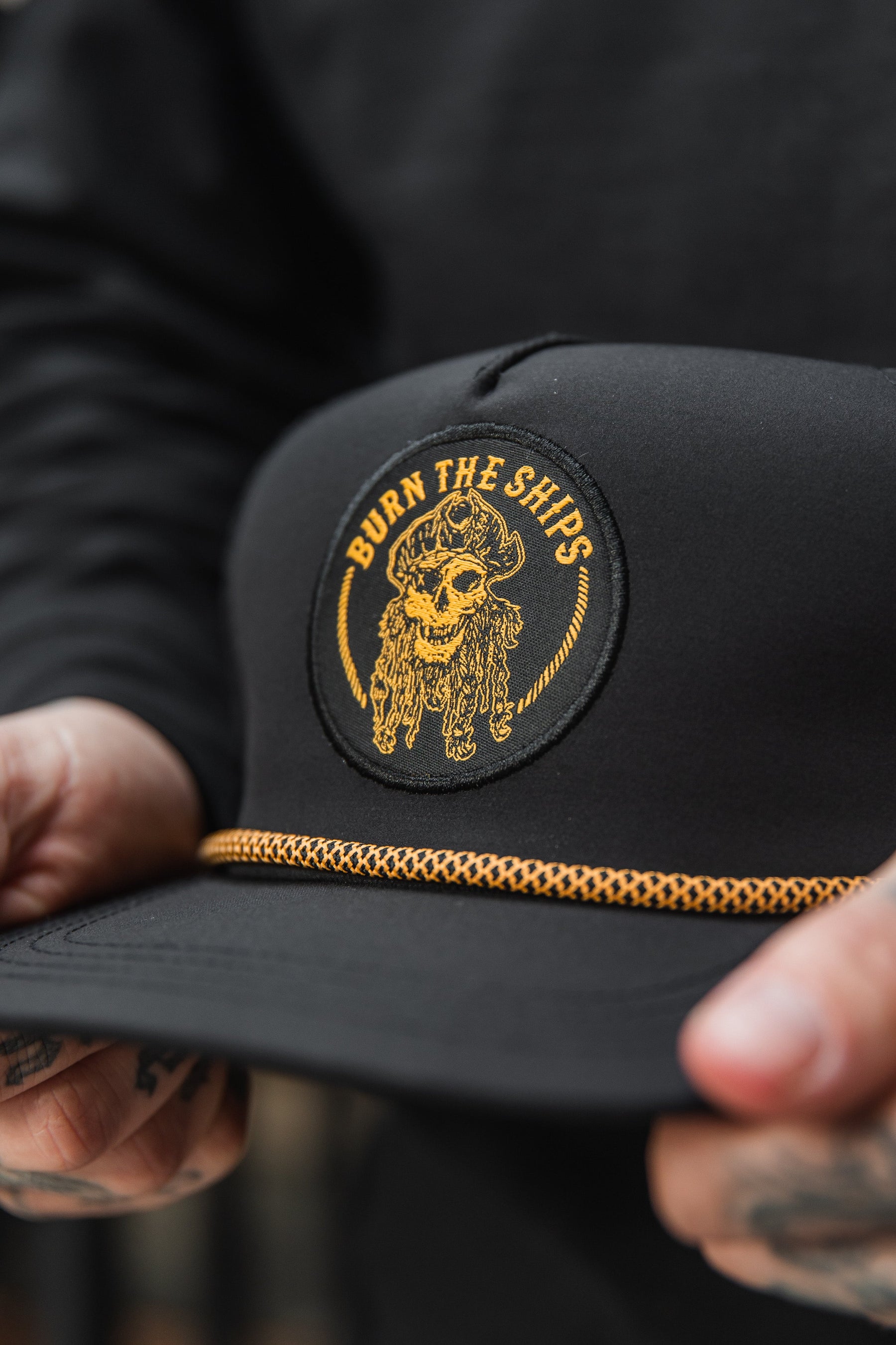 Close-up of a person holding the Relentless Betrayal Burn The Ships BLACKOUT Snapback, featuring a skull pirate patch with Burn the Ships in yellow. This handmade hat includes a decorative yellow rope above the brim, while tattoos adorn the persons fingers.